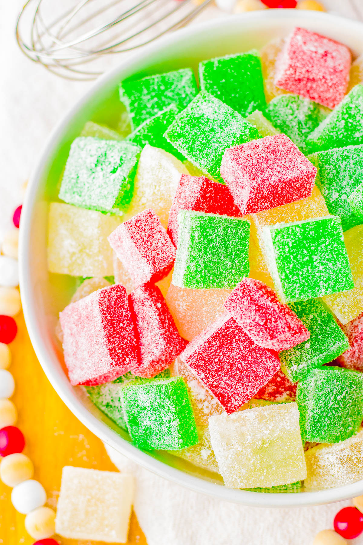 A bowl filled with colorful sugar-coated jelly cubes in green, red, and yellow. A mixing whisk and beaded decoration are partially visible around the bowl.