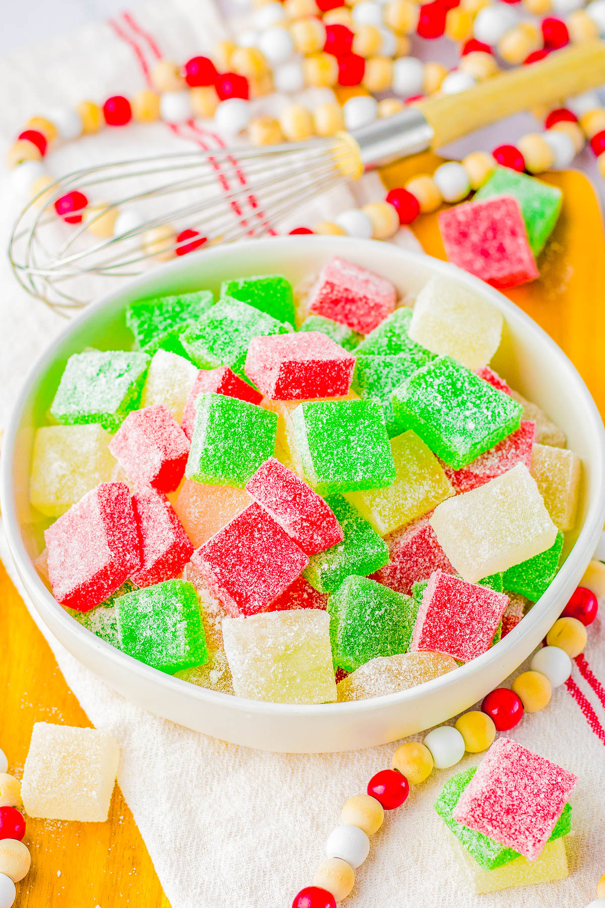 A bowl of red, green, and white sugar-coated gummy candies on a wooden board with a whisk and beaded garland in the background.