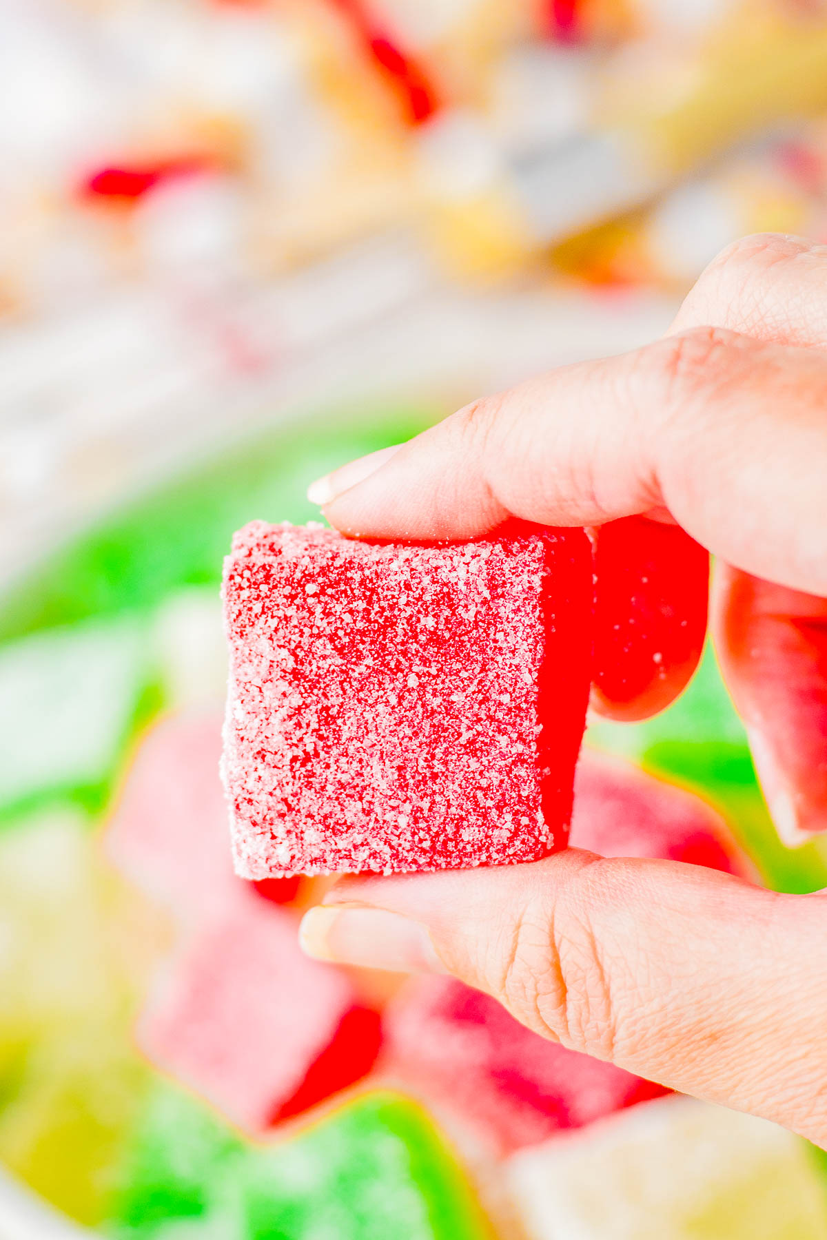 A hand holding a sugar-coated red gummy candy with other colorful candies blurred in the background.