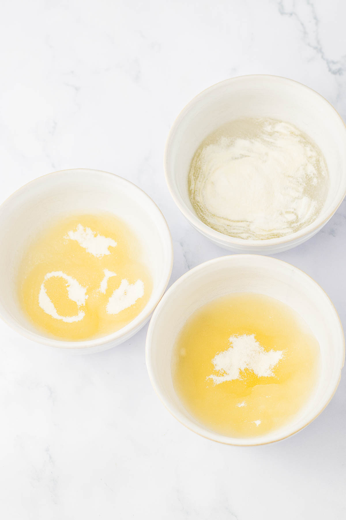 Three white bowls on a marble surface containing small amounts of yellow liquid with white powder sprinkled on top.