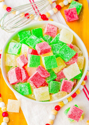 A bowl of colorful, sugar-coated gumdrops in red, green, and yellow on a wooden tray with a whisk nearby.