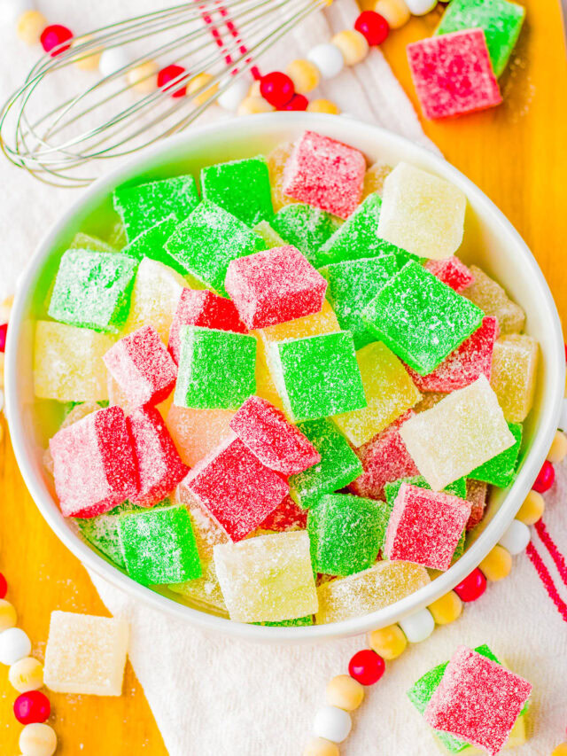 A bowl of colorful, sugar-coated gumdrops in red, green, and yellow on a wooden tray with a whisk nearby.