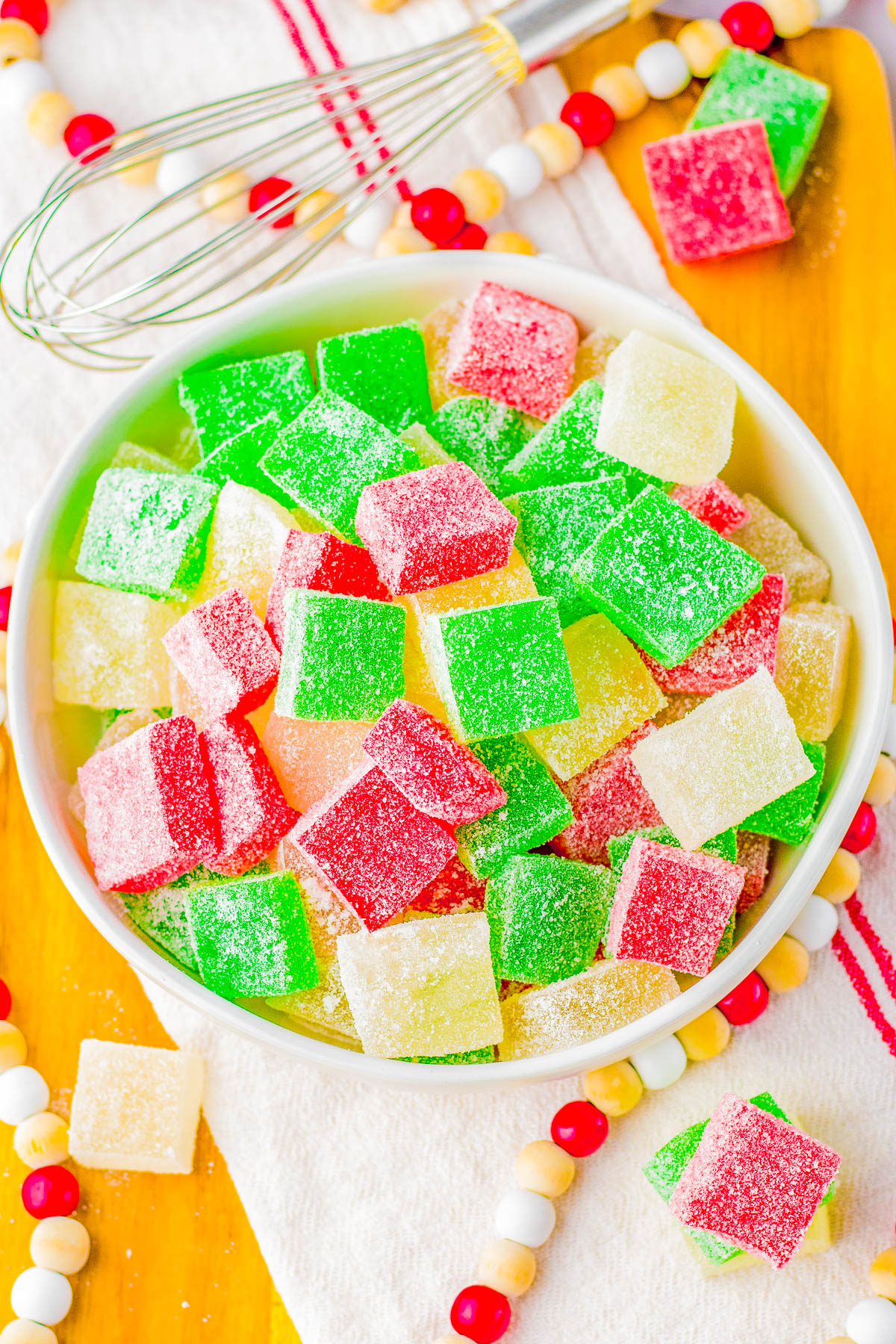 A bowl of colorful, sugar-coated gummy cubes in red, green, and yellow on a wooden tray with a whisk nearby.