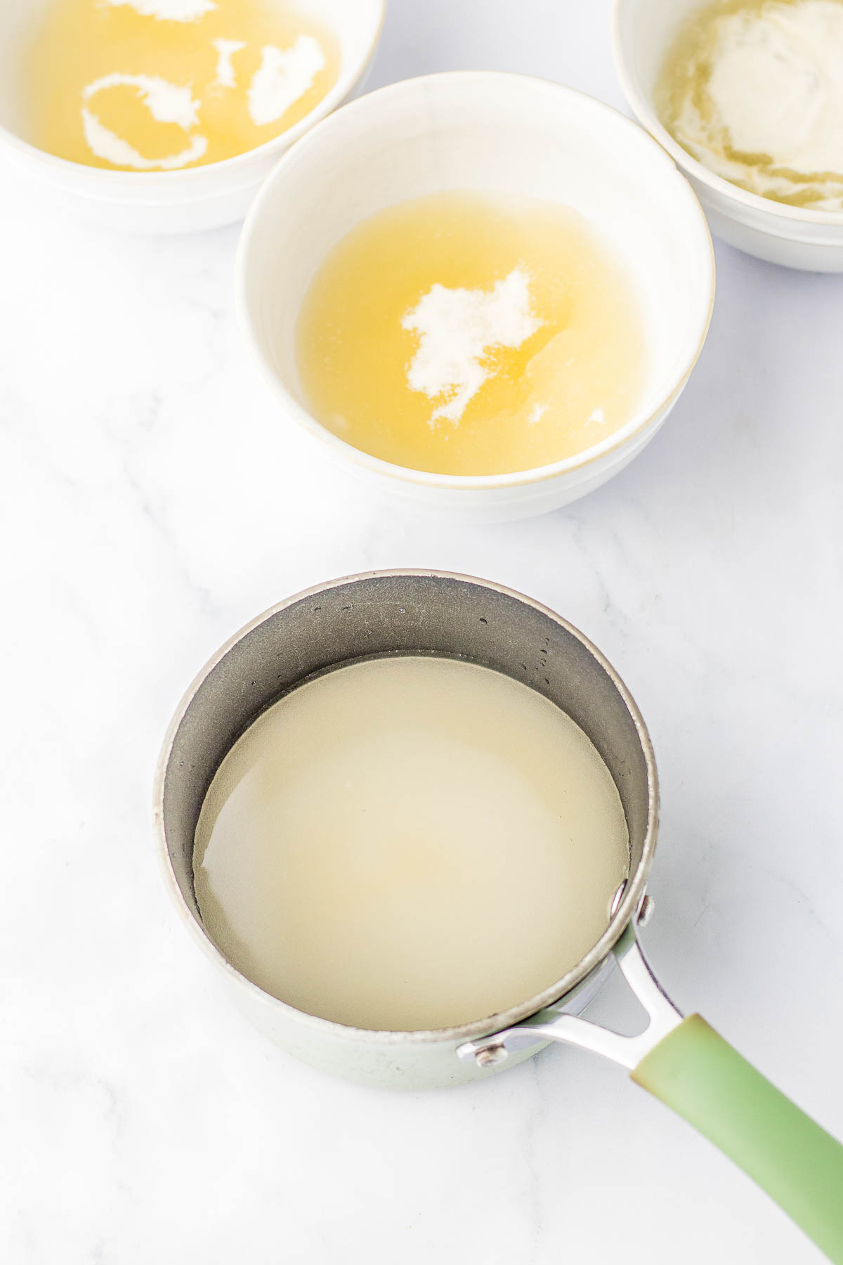 Small saucepan with liquid and three bowls containing melted butter and other ingredients on a marble countertop.