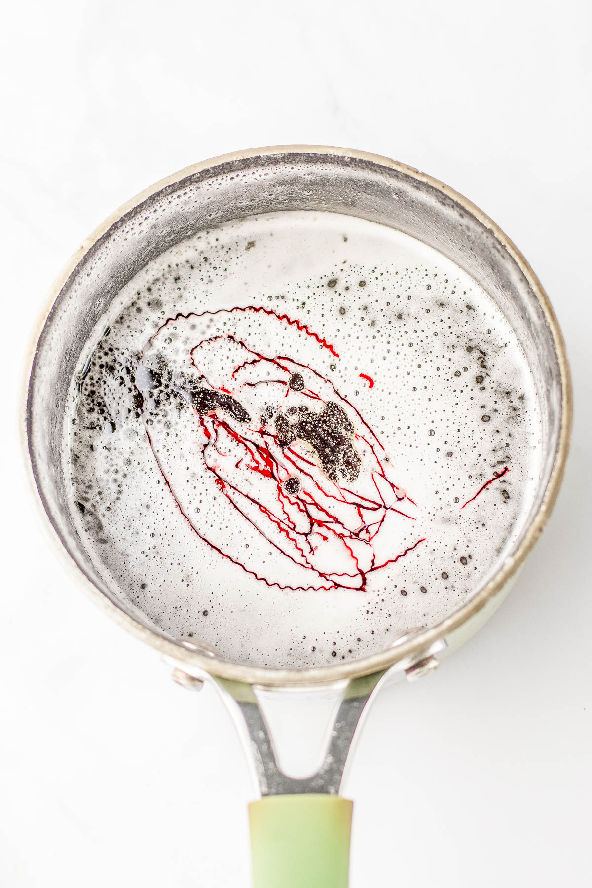 A saucepan contains frothy liquid with a drizzle of red dye forming abstract patterns on the surface.