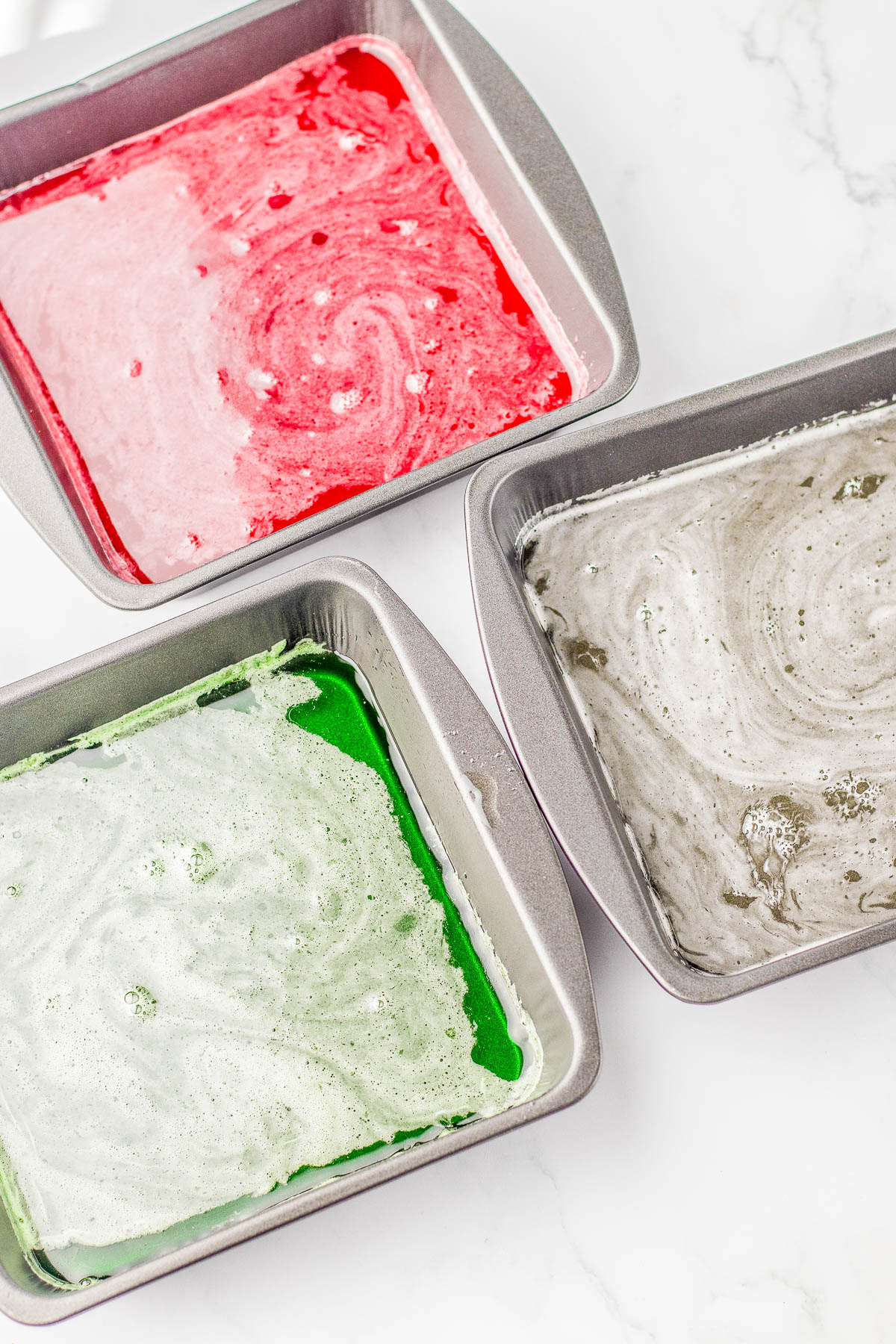 Three square baking pans filled with red, green, and gray swirled batter on a white surface.