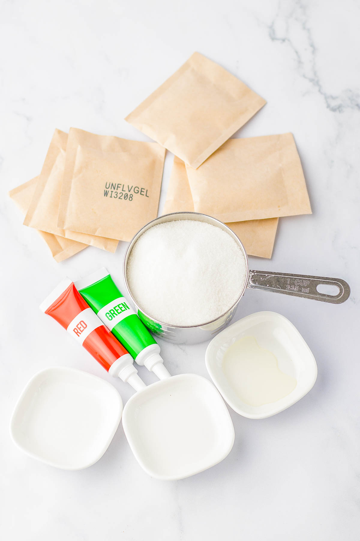 Packets of unflavored gelatin, a measuring cup of sugar, red and green food coloring tubes, and two small bowls on a marble surface.