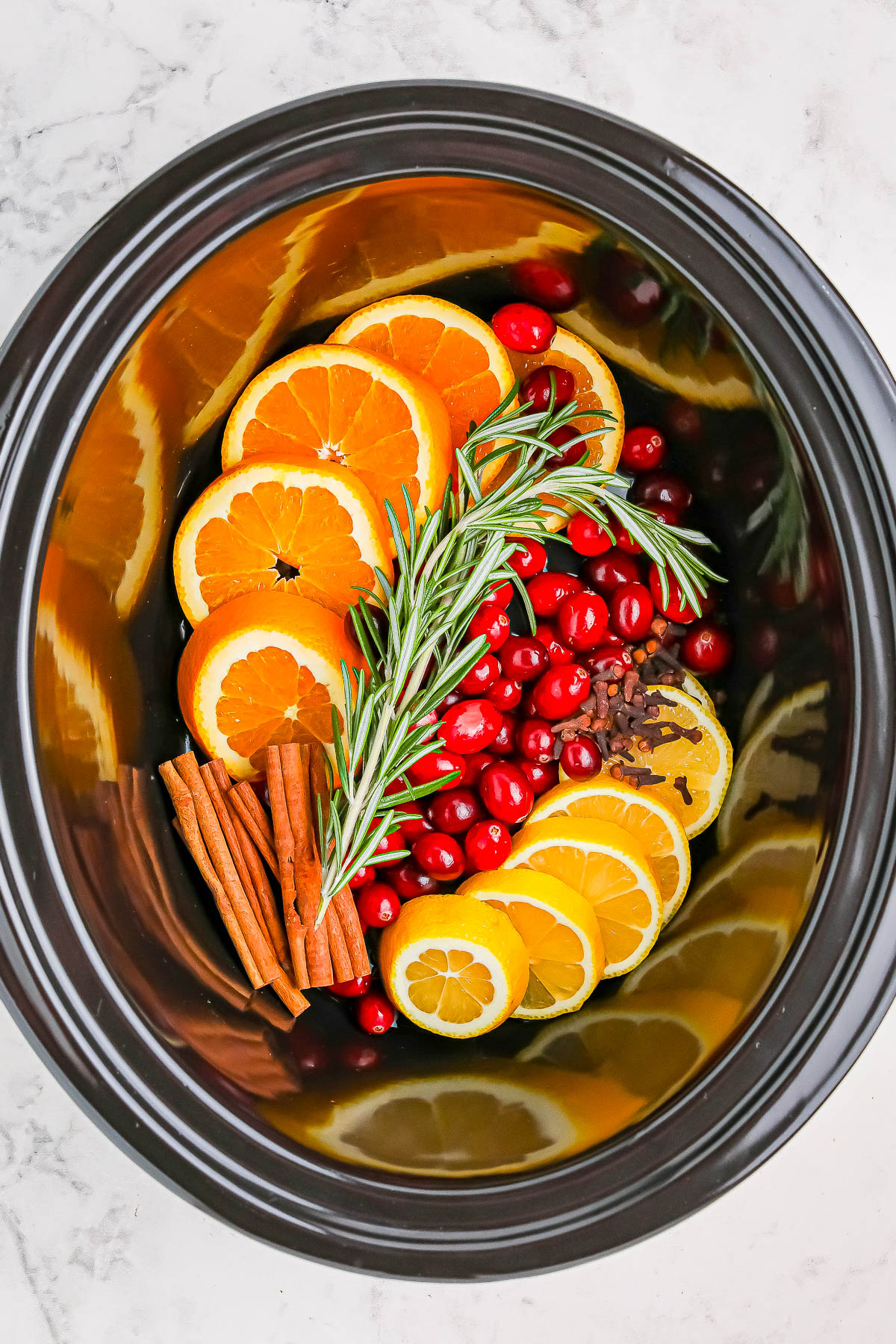 Sliced oranges, lemons, cranberries, cinnamon sticks, and rosemary in a slow cooker.