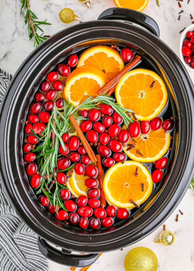 A slow cooker filled with sliced oranges, cranberries, cinnamon sticks, rosemary, and cloves on a marble surface.