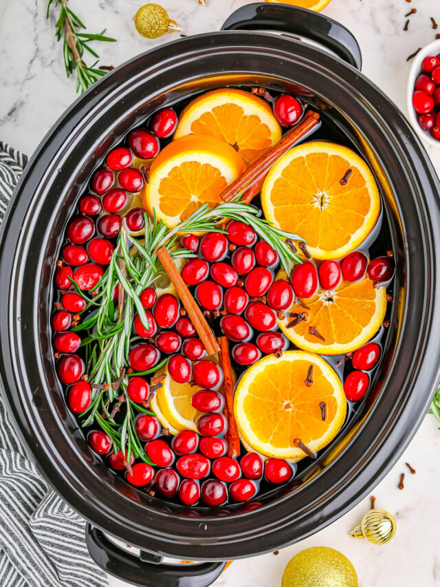 A slow cooker filled with sliced oranges, cranberries, cinnamon sticks, rosemary, and cloves on a marble surface.