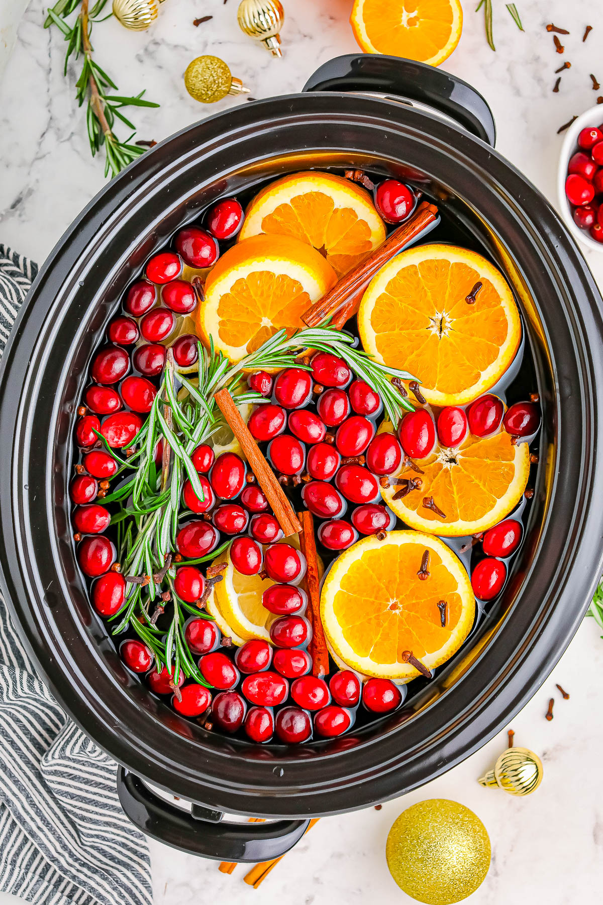 A slow cooker filled with sliced oranges, cranberries, cinnamon sticks, rosemary, and cloves on a marble surface.