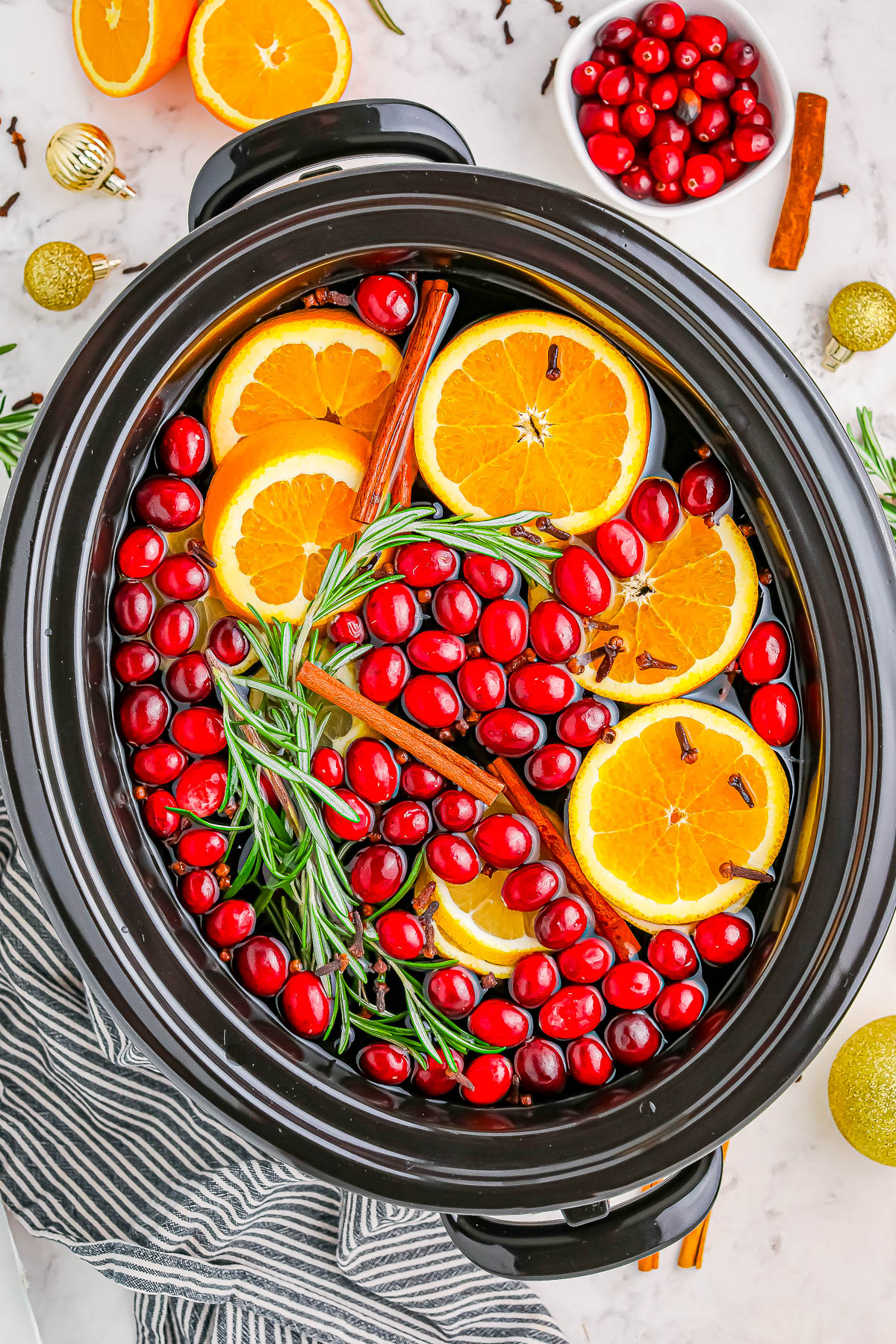 A slow cooker filled with sliced oranges, cranberries, rosemary, cinnamon sticks, and cloves.