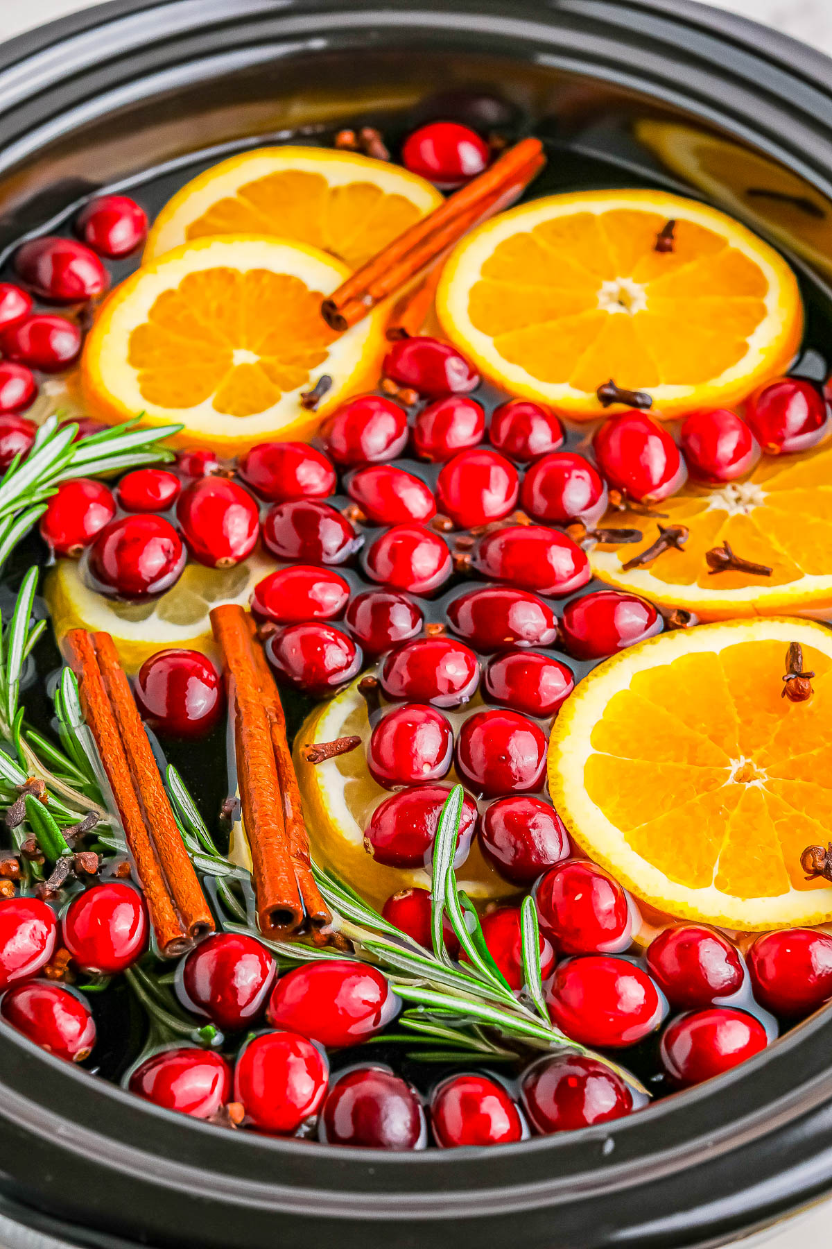 A pot filled with water contains floating orange slices, cranberries, cinnamon sticks, rosemary sprigs, and cloves.