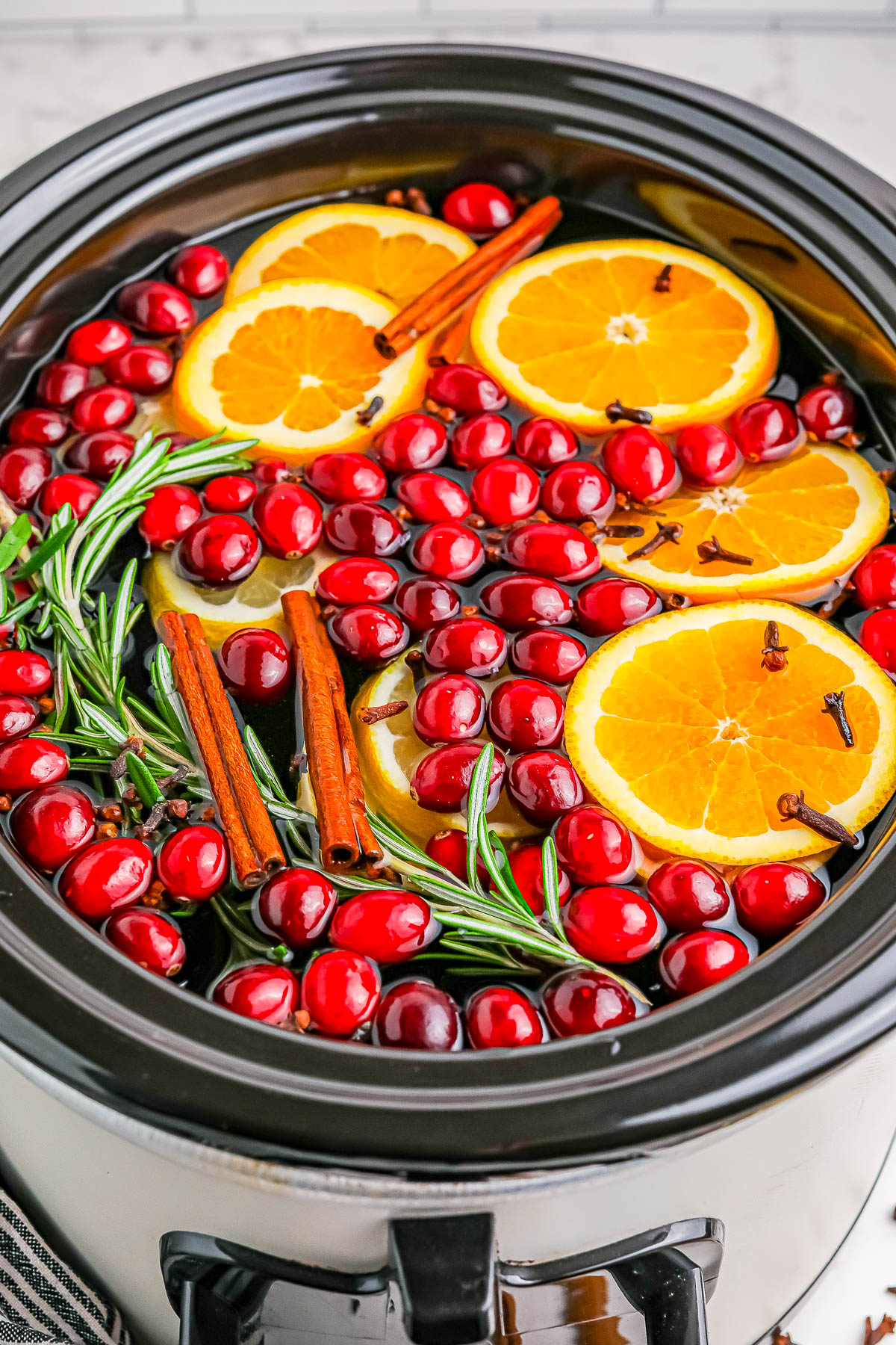 Slow cooker filled with cranberries, orange slices, rosemary, cinnamon sticks, and cloves.