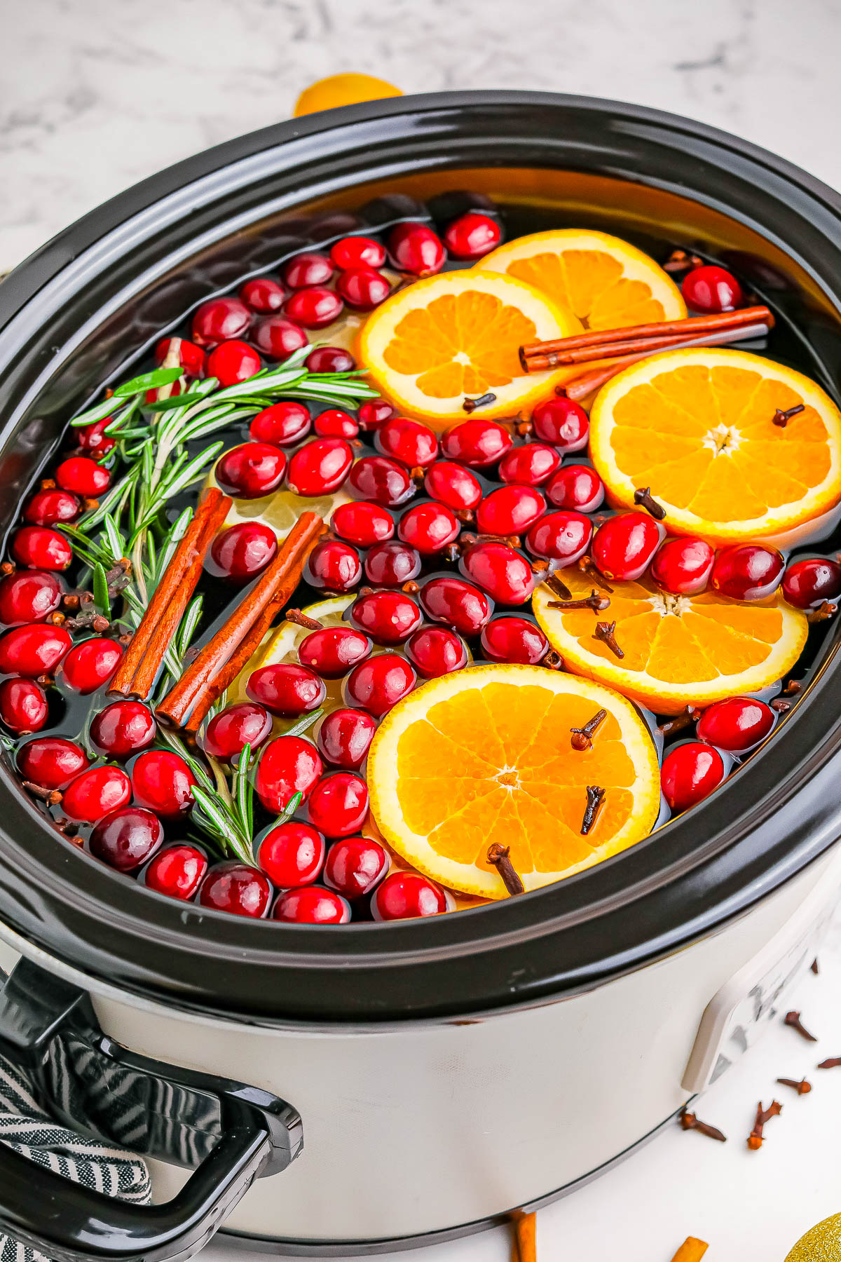 A slow cooker filled with water, fresh cranberries, orange slices, cinnamon sticks, rosemary sprigs, and cloves on a marble countertop.