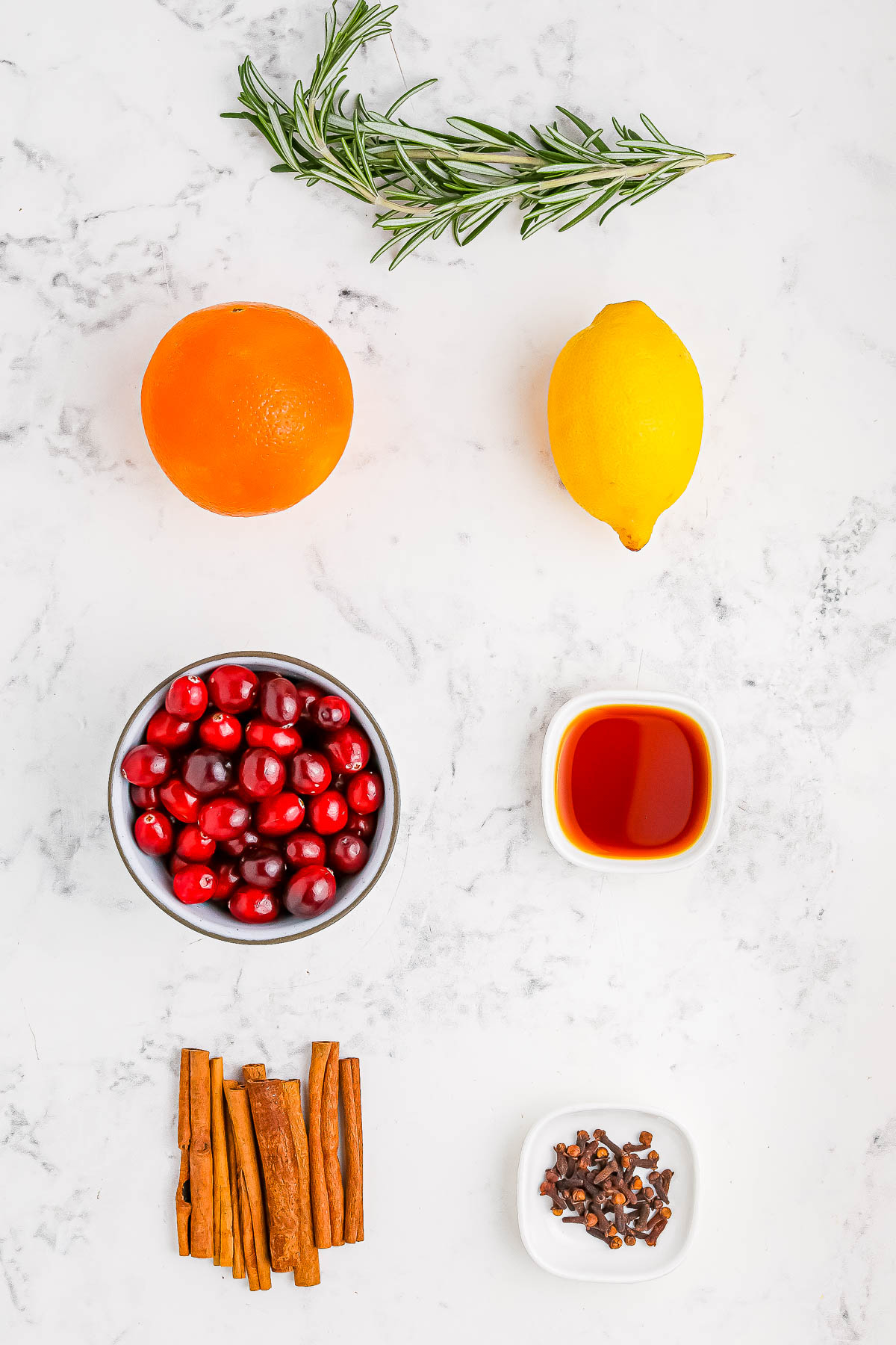 Ingredients for a recipe: a sprig of rosemary, an orange, a lemon, a bowl of cranberries, cinnamon sticks, cloves, and a small bowl of liquid on a marble surface.