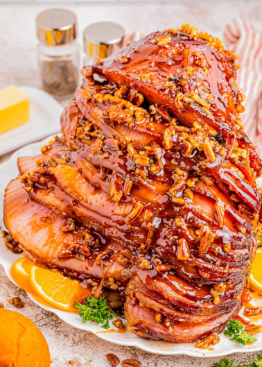 A spiral-cut ham glazed with honey and pecan topping, garnished with orange slices and parsley on a white plate. Butter and condiments are in the background.