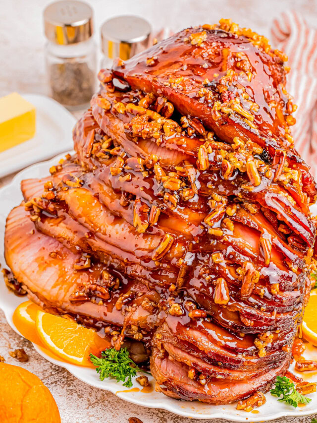 A spiral-cut ham glazed with honey and pecan topping, garnished with orange slices and parsley on a white plate. Butter and condiments are in the background.