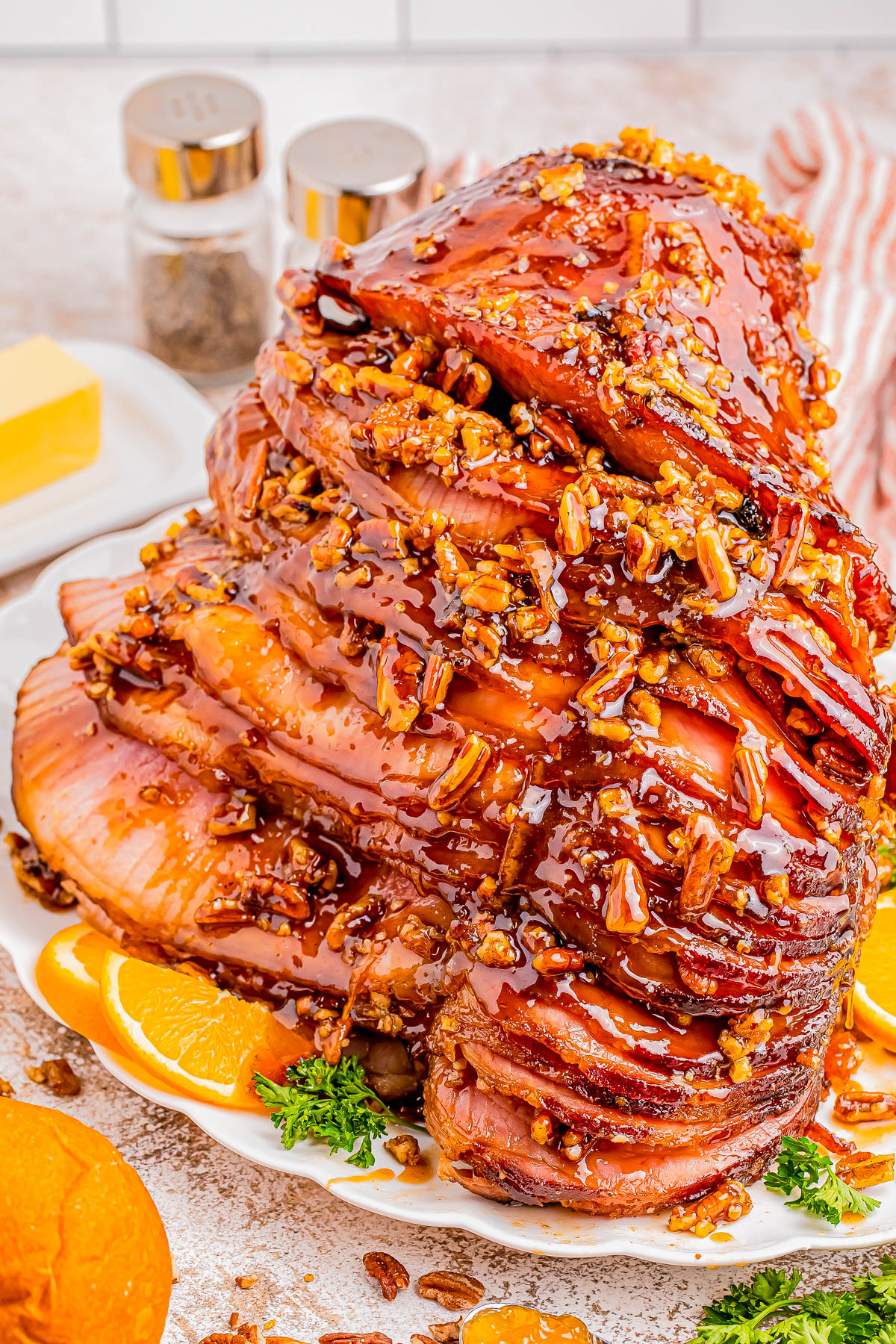 A spiral-cut ham glazed with honey and pecan topping, garnished with orange slices and parsley on a white plate. Butter and condiments are in the background.