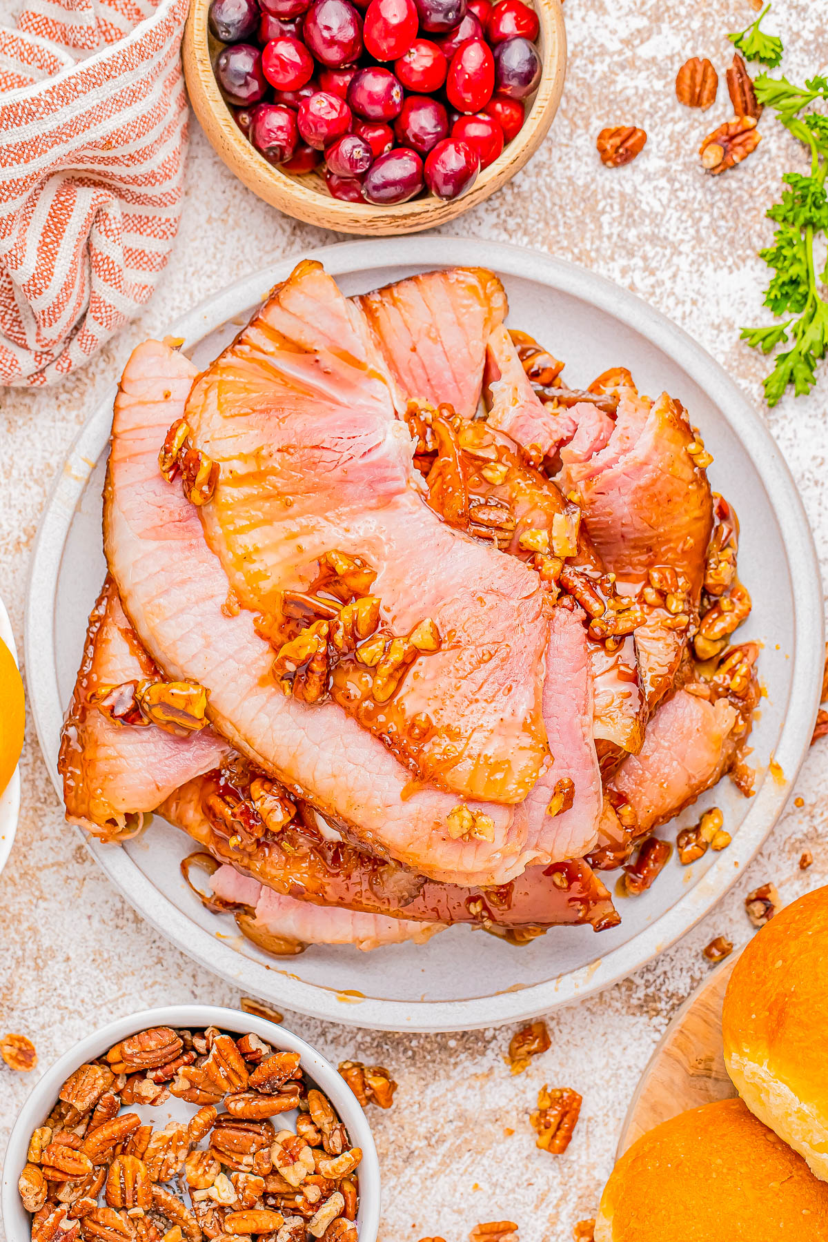 A plate of glazed ham slices topped with pecans, surrounded by cranberries, pecans, bread rolls, and a cloth napkin on a textured surface.