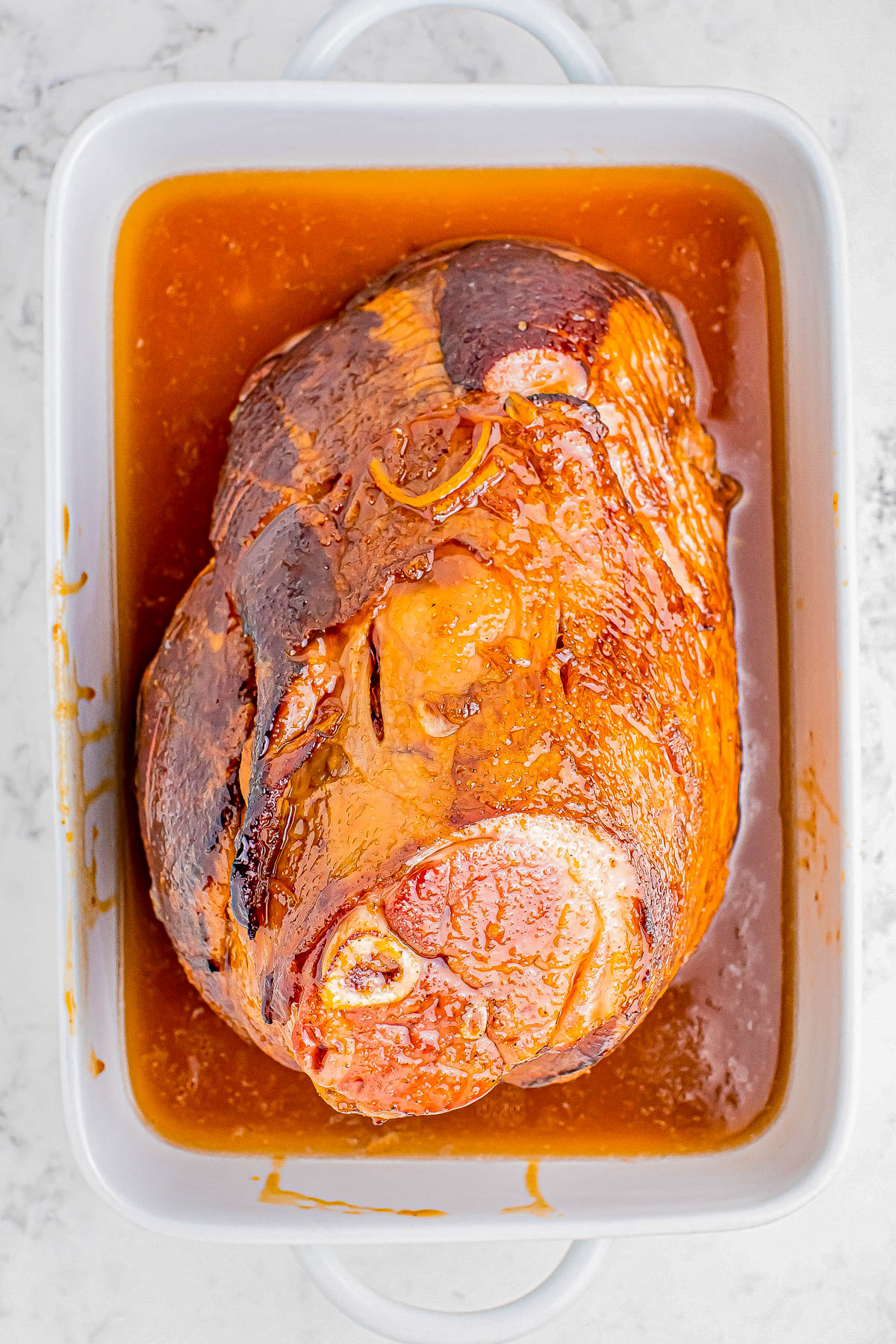 Cooked ham in a white roasting pan with brown glaze, placed on a marble surface.