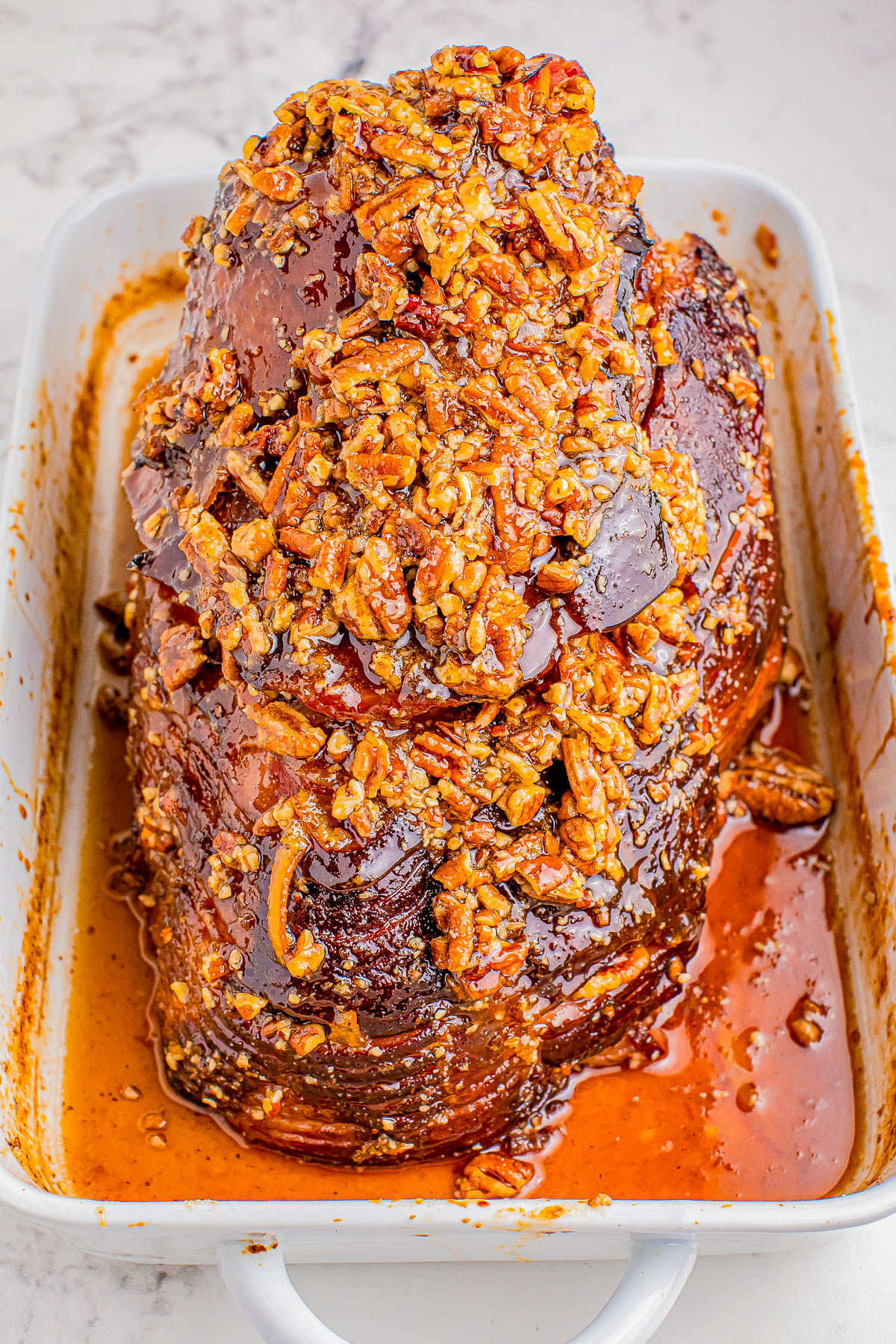 Cooked ham topped with a brown sugar and nut glaze, placed in a white baking dish on a marble surface.