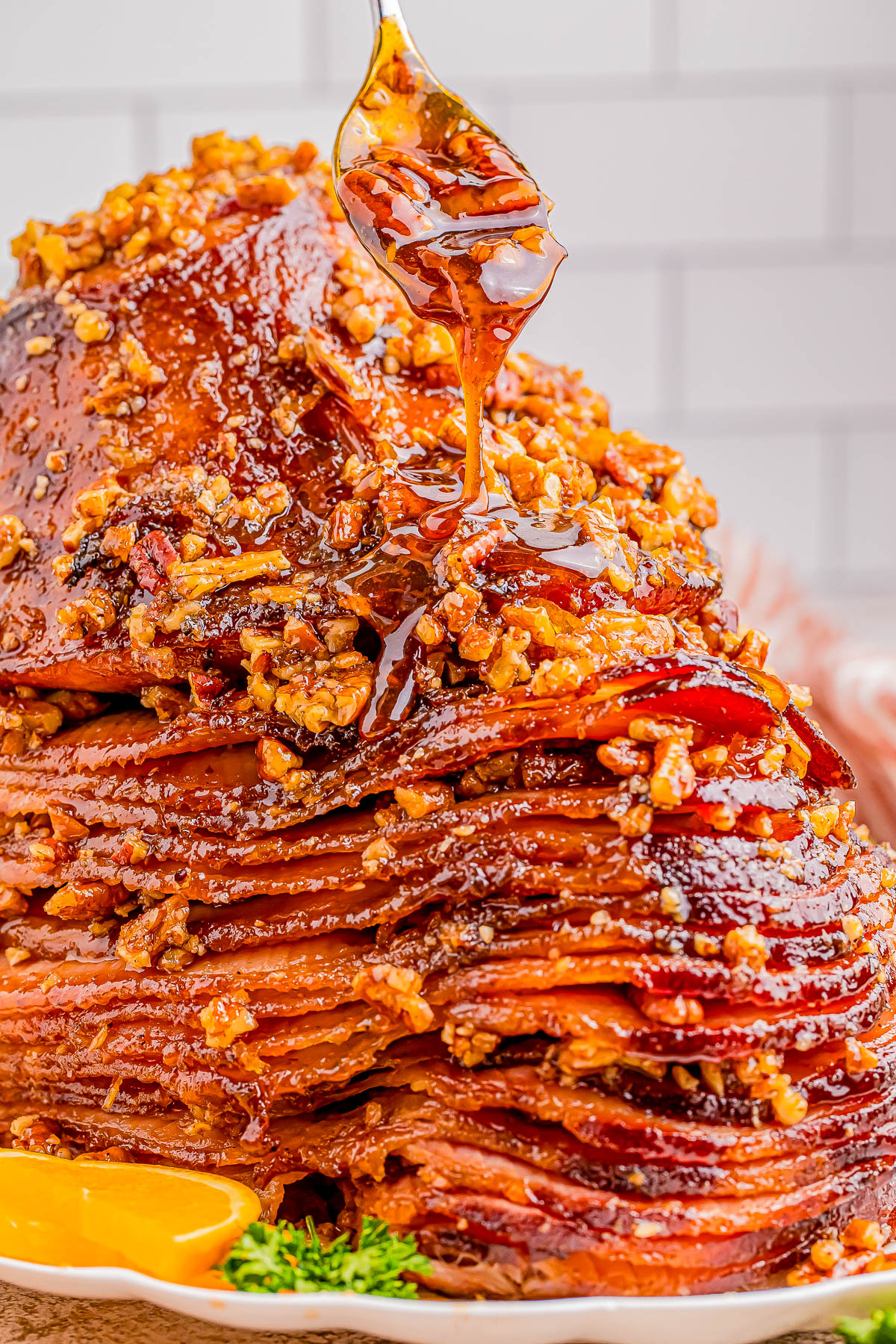 A spoon drizzles glaze over a stack of sliced ham, topped with a nutty crust, served on a white plate with parsley garnish.