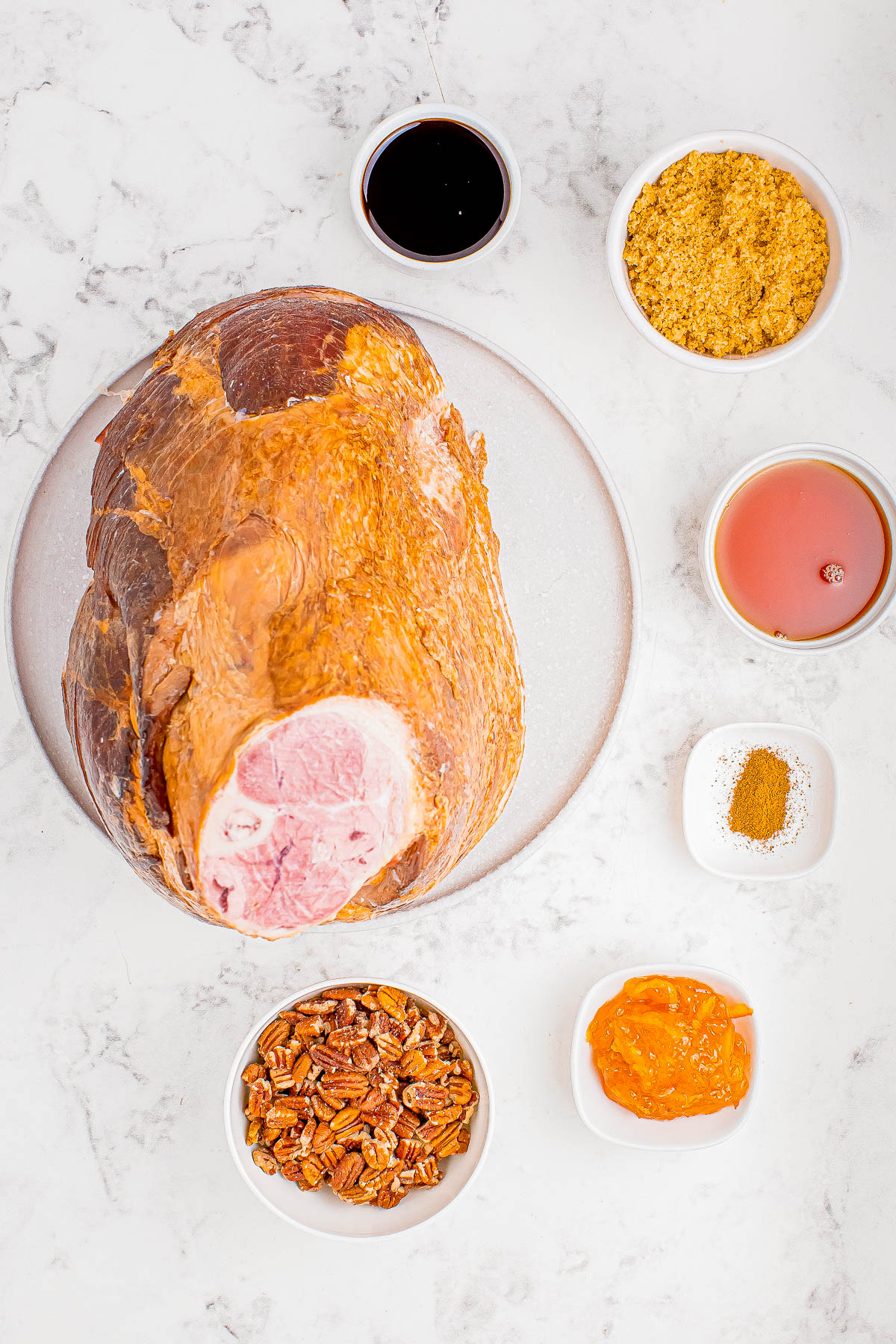 A large ham on a plate surrounded by small bowls containing soy sauce, brown sugar, apple cider, spices, pecans, and apricot jam on a marble surface.