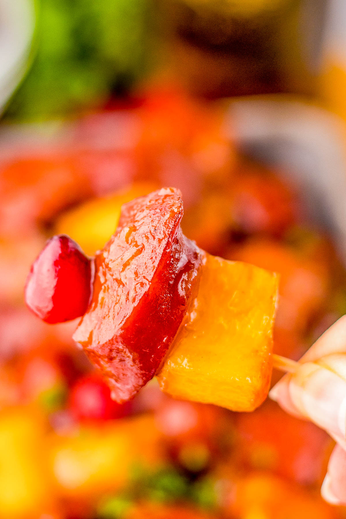 Close-up of a skewer with glazed ham cubes and pineapple chunks, with a blurred colorful background.