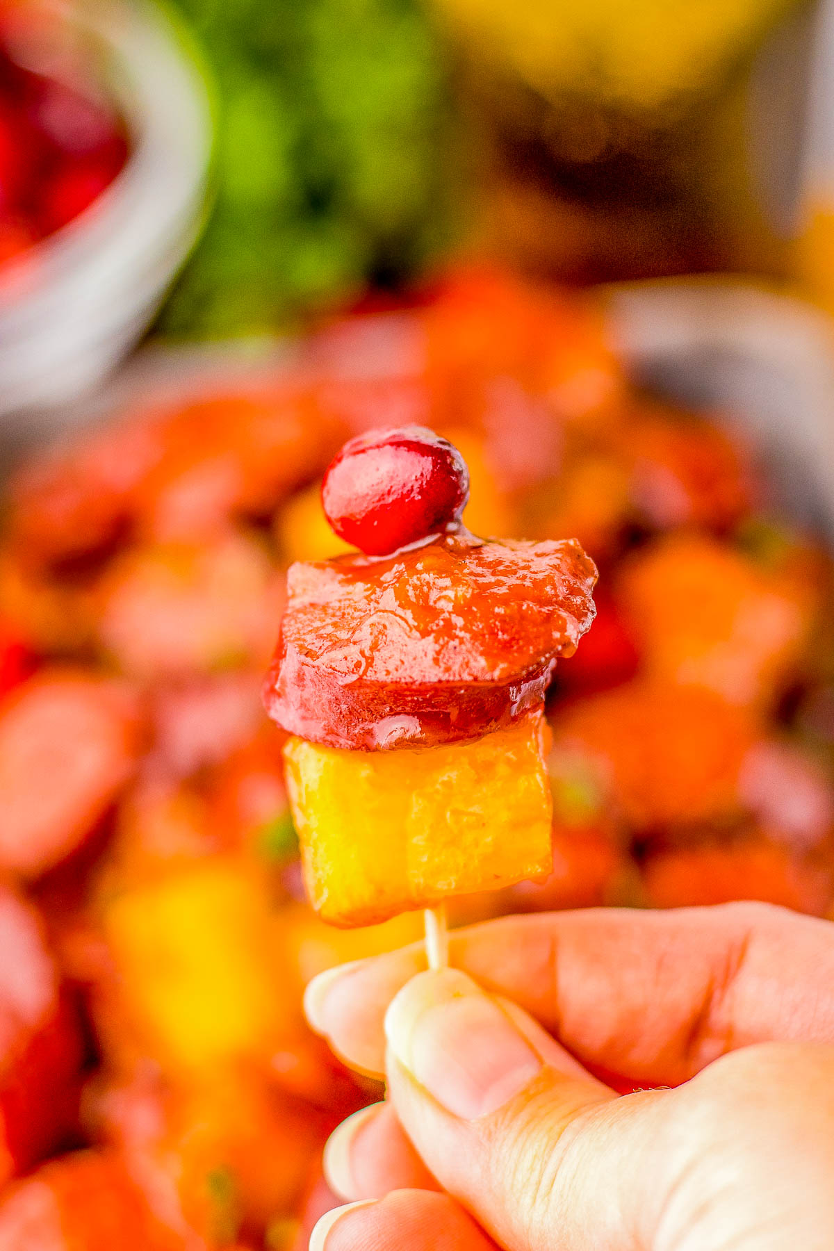 A hand holds a skewer with pineapple, sausage, and a cherry on top against a blurred colorful background.