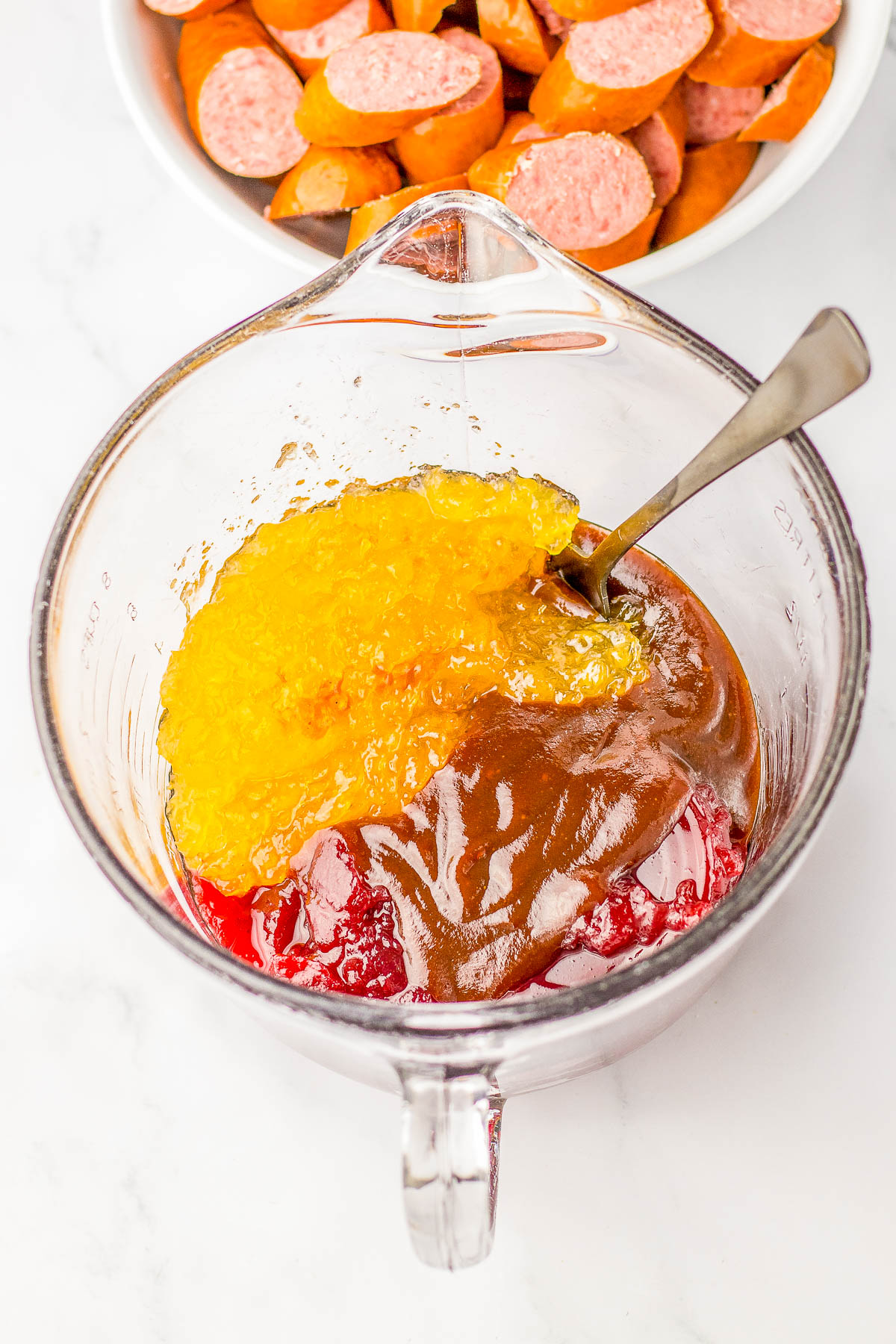 A bowl with a mixture of red and brown sauces topped with a yellow jelly, beside a fork and a bowl of sliced sausages.