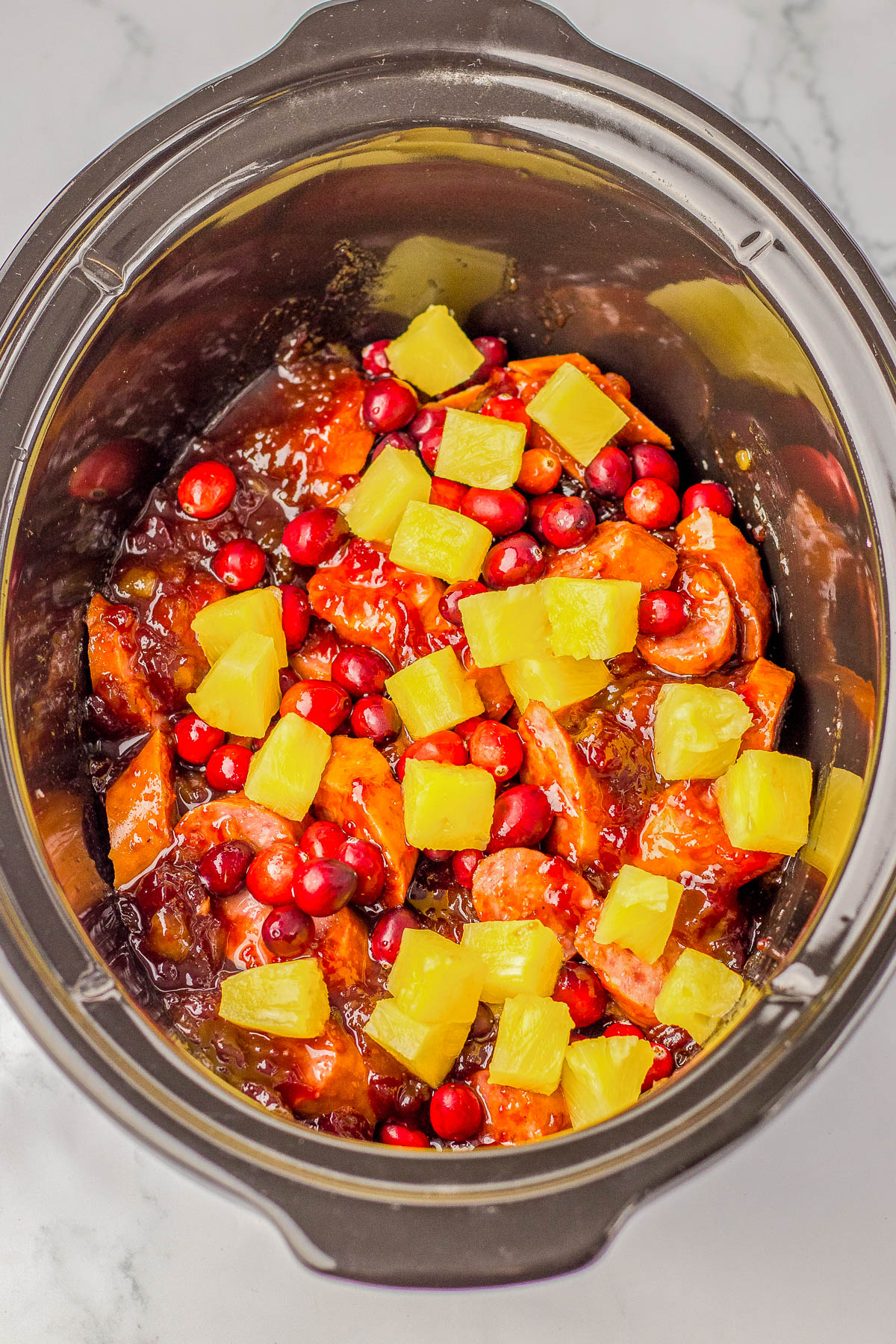 Slow cooker containing a dish with sliced sausages, cranberries, pineapple chunks, and sauce.