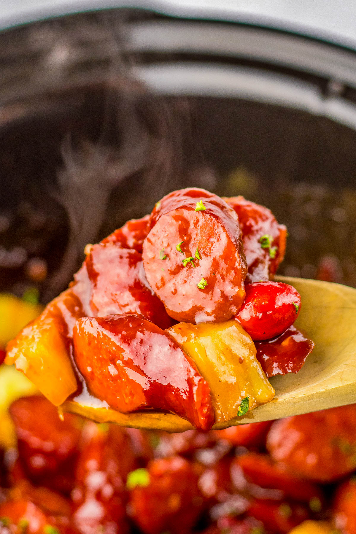 Close-up of a wooden spoon holding steaming pieces of sausage and pineapple glazed in a red sauce, with more ingredients visible in the background.