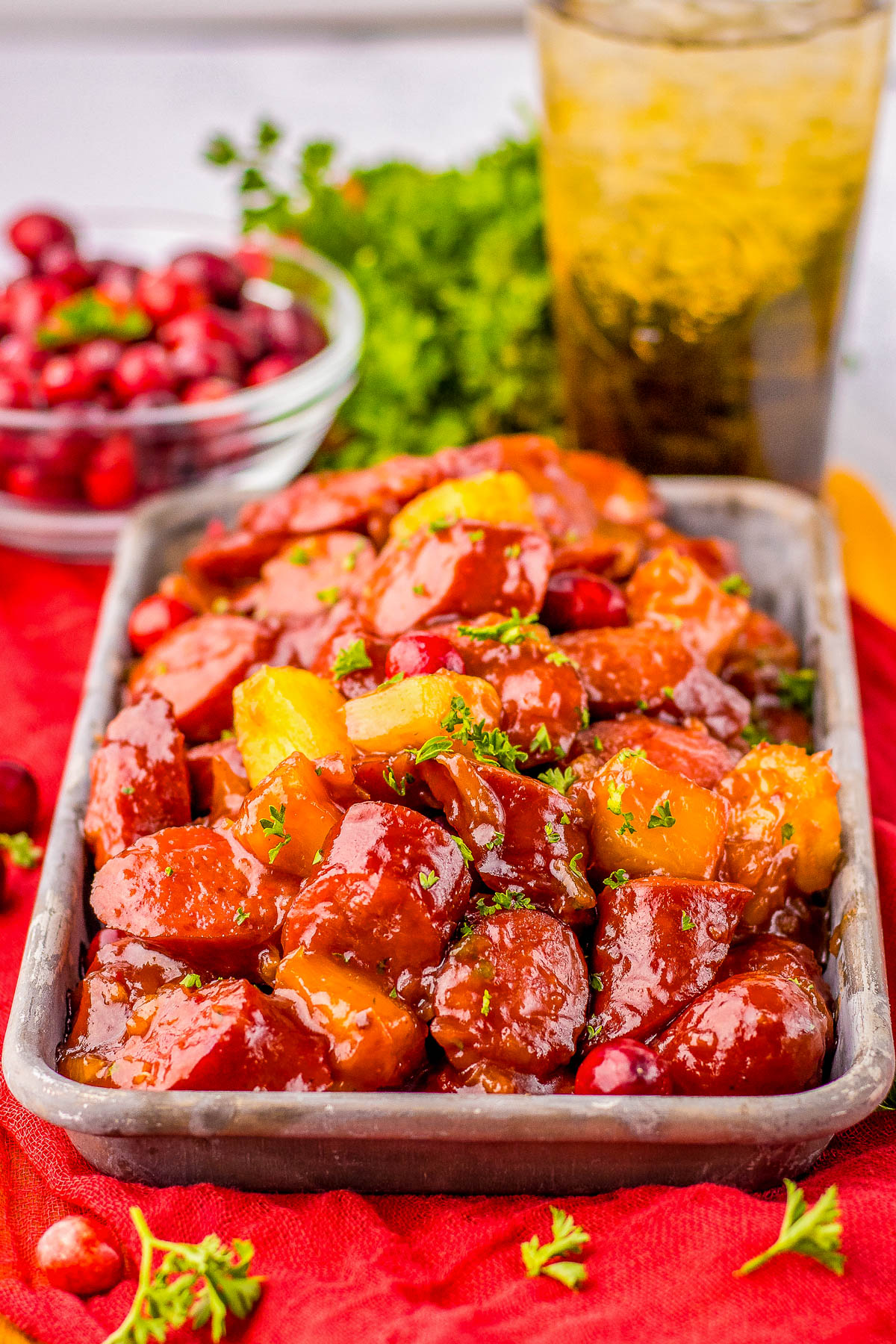 A tray of glazed sausage pieces and pineapple chunks garnished with herbs. A bowl of cranberries and a glass of iced drink are in the background.