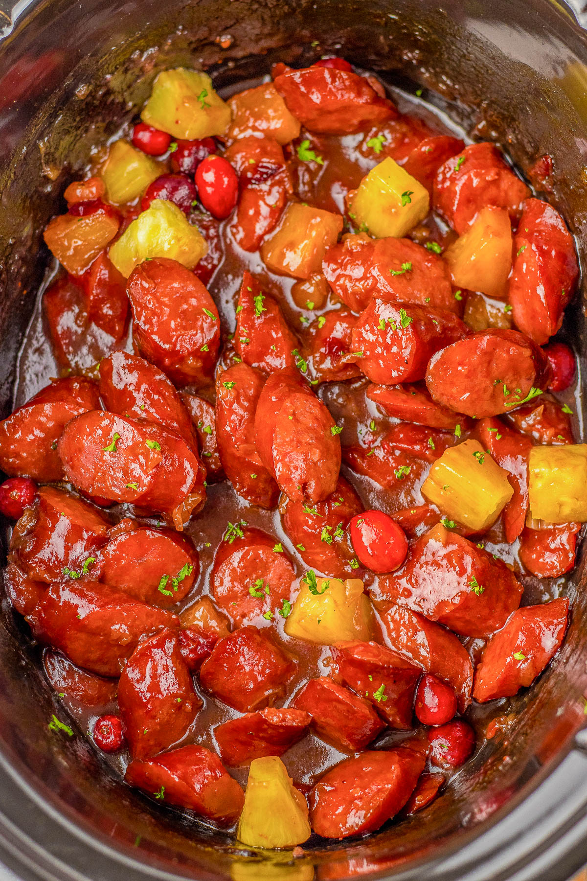 Slow-cooked dish with sliced sausages, pineapple chunks, and cranberries in a glossy sauce.