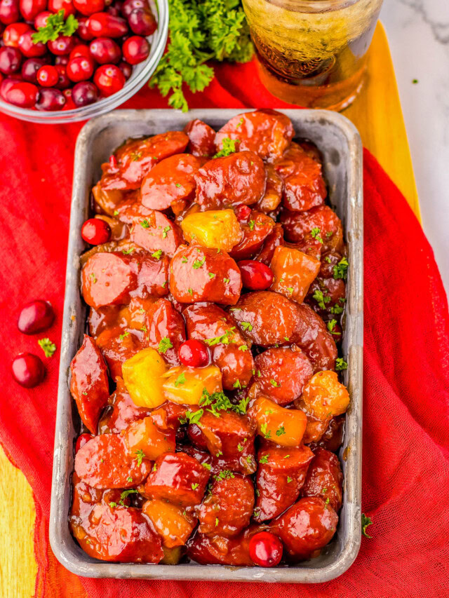 Rectangular dish with glazed sausage slices and pineapple chunks, garnished with parsley. A bowl of cranberries and a glass of iced drink are in the background, on a red cloth.