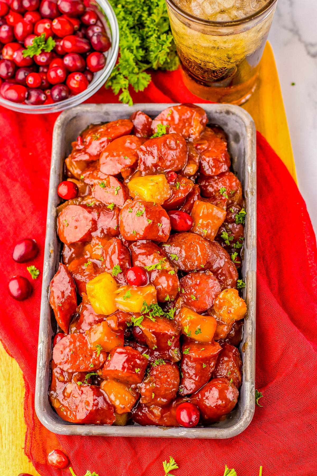 Rectangular dish with glazed sausage slices and pineapple chunks, garnished with parsley. A bowl of cranberries and a glass of iced drink are in the background, on a red cloth.