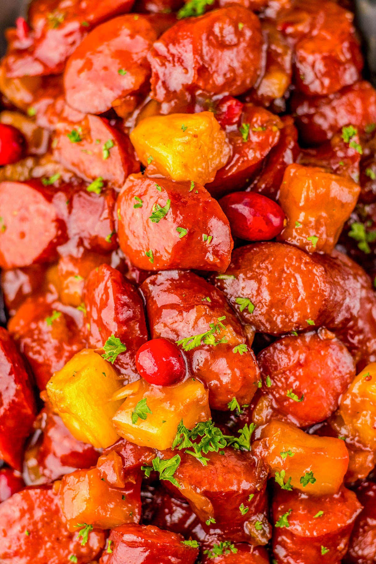 Close-up of glazed sausage pieces mixed with pineapple chunks and cranberries, garnished with chopped herbs.