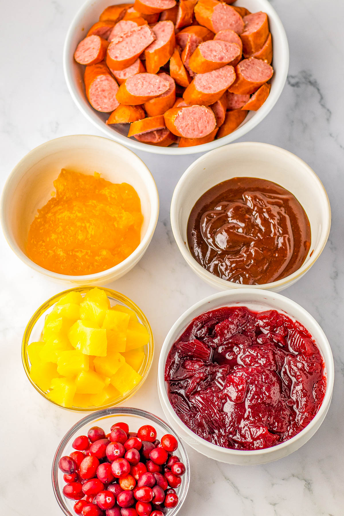 Bowls of sliced sausage, orange jelly, brown sauce, diced pineapple, red sauce, and cranberries on a marble surface.