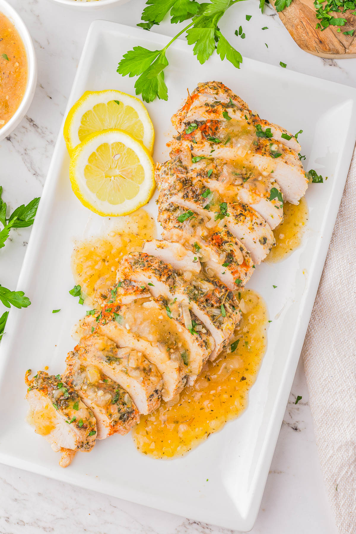 Sliced grilled chicken breast with herb seasoning topped with sauce, garnished with lemon slices and parsley on a white rectangular plate.