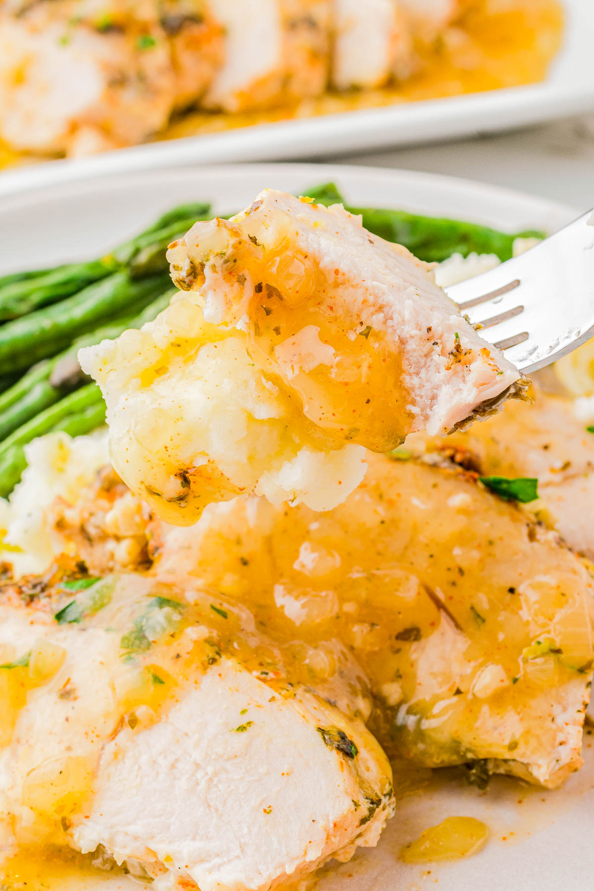 A fork holds a piece of chicken with gravy over mashed potatoes and asparagus on a plate.