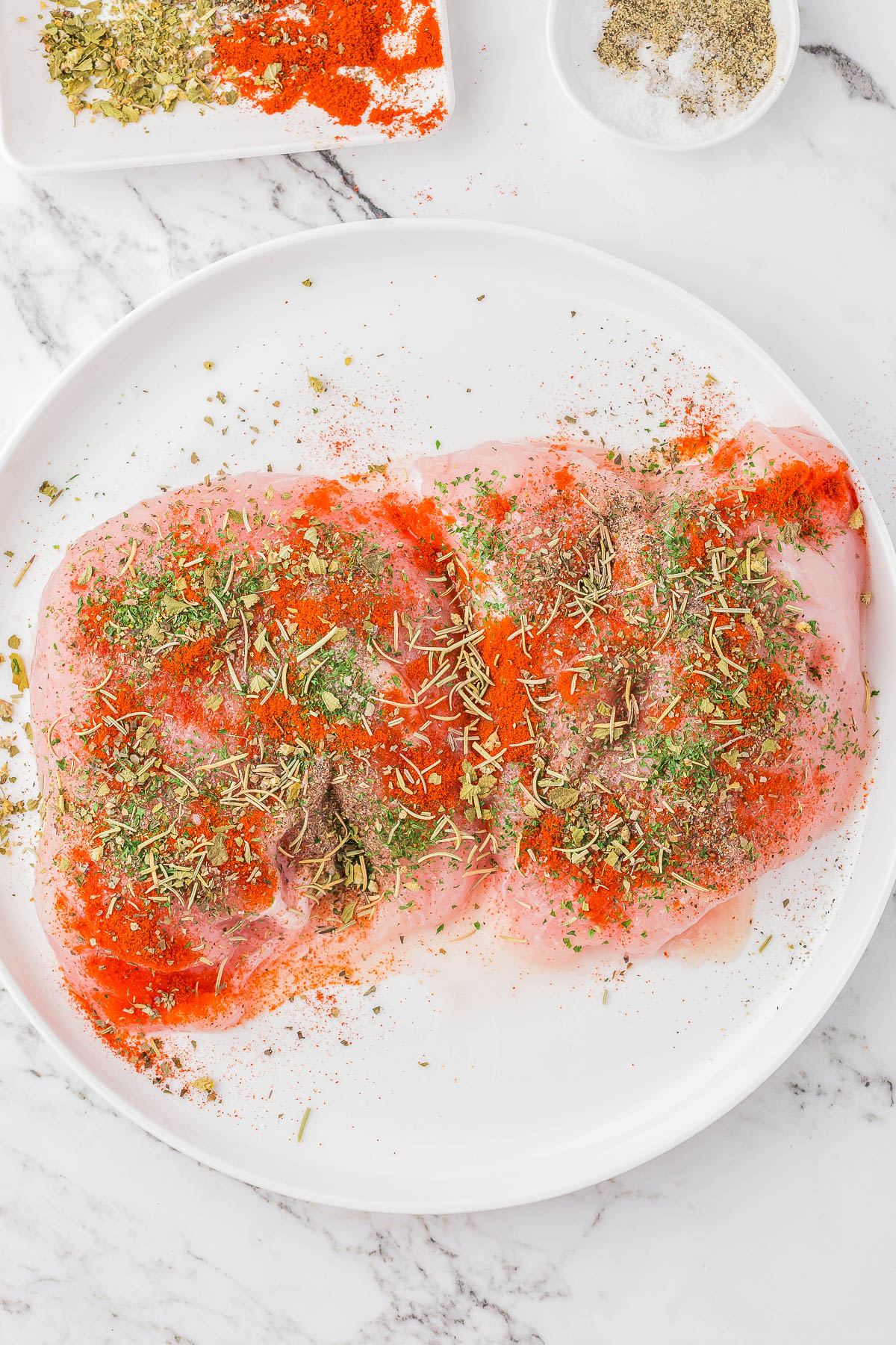 Raw chicken breast seasoned with various herbs and spices on a white plate.