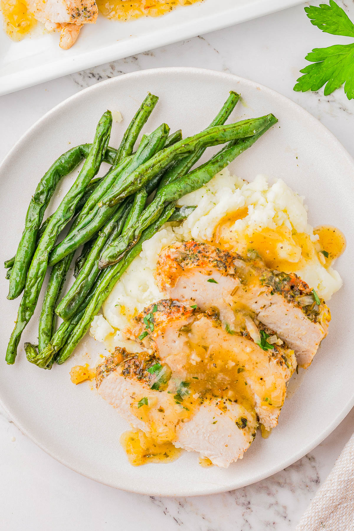 Plate with sliced chicken breast in herb sauce, mashed potatoes, and green beans on a white plate.