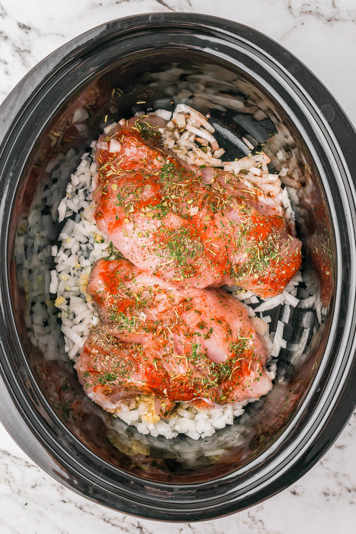 Raw seasoned chicken breasts on a bed of chopped onions in a slow cooker.