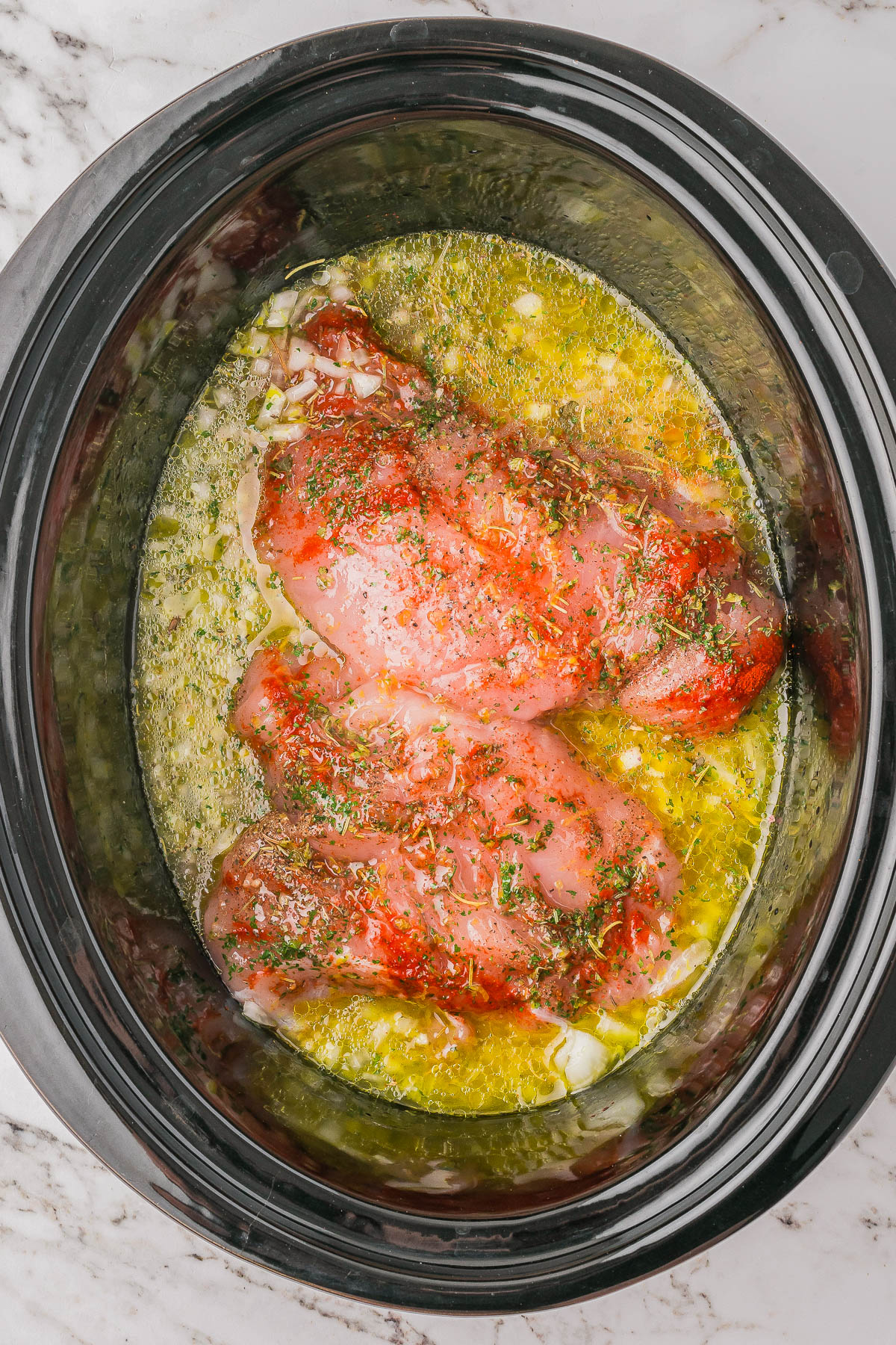 Raw chicken breasts seasoned with herbs and spices in a slow cooker with broth.