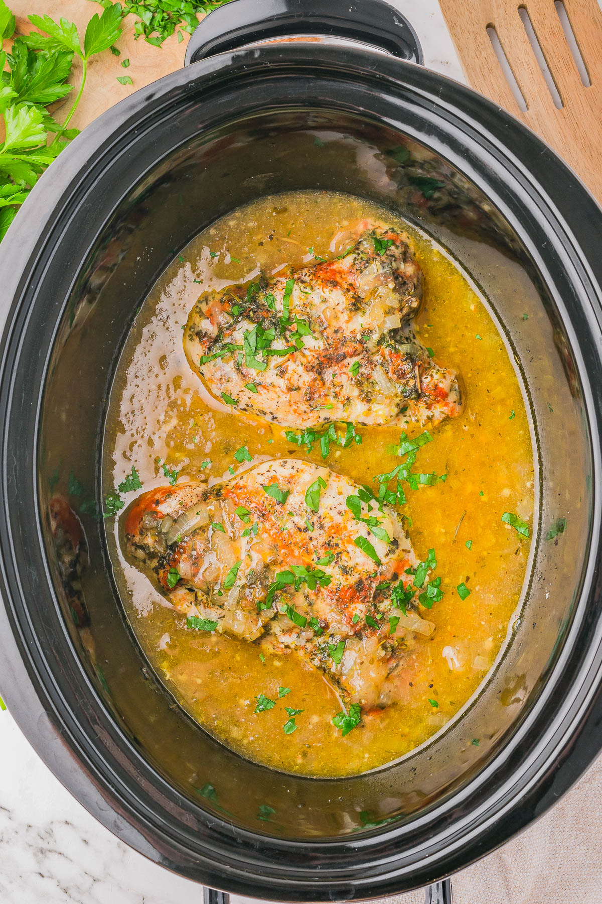 Two seasoned chicken breasts cooked in a slow cooker with brothy sauce, garnished with chopped parsley.