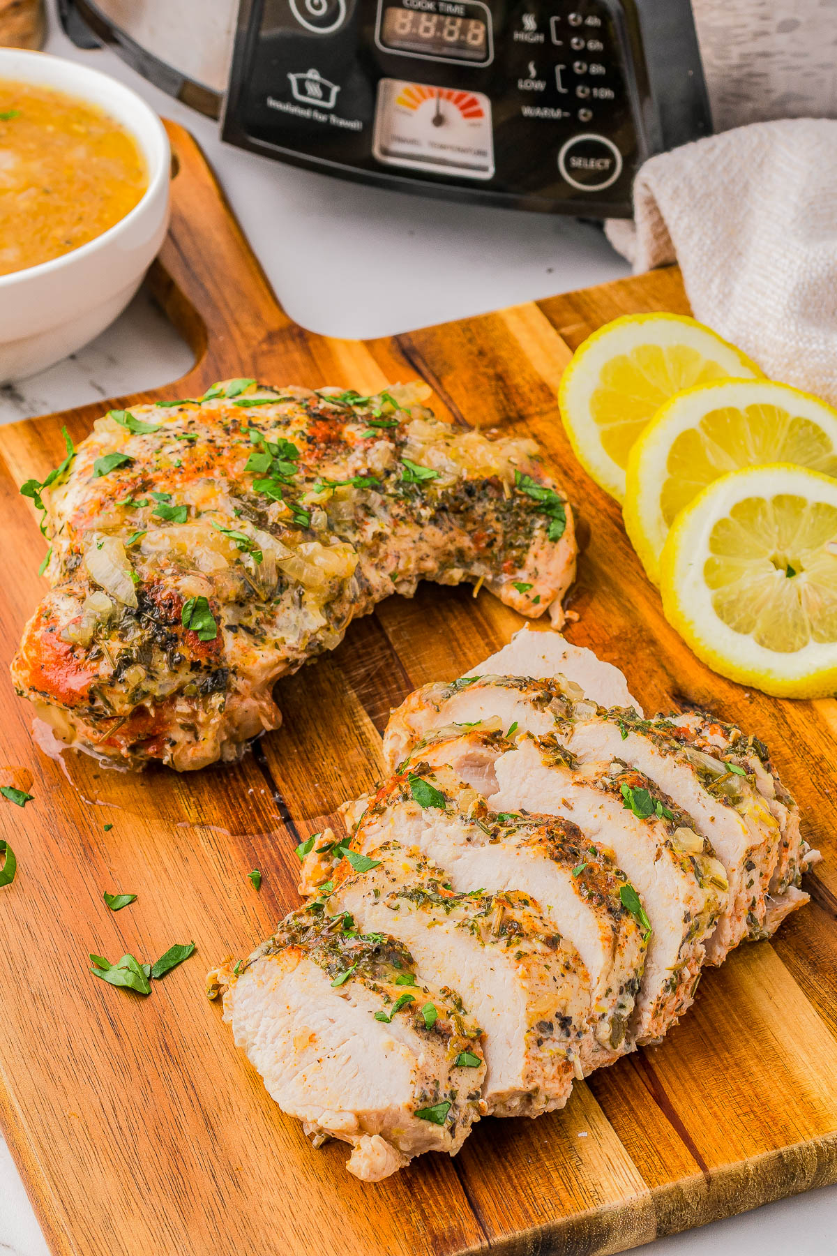 Sliced and unsliced seasoned chicken breasts on a wooden board, garnished with herbs, alongside lemon slices and a bowl of sauce, with a kitchen appliance in the background.