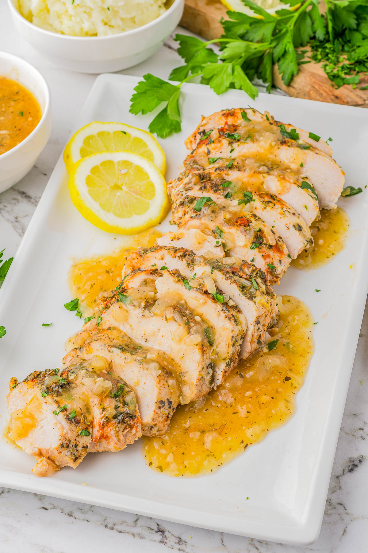 Sliced cooked chicken breast topped with a light sauce, garnished with parsley and lemon slices, on a white plate.