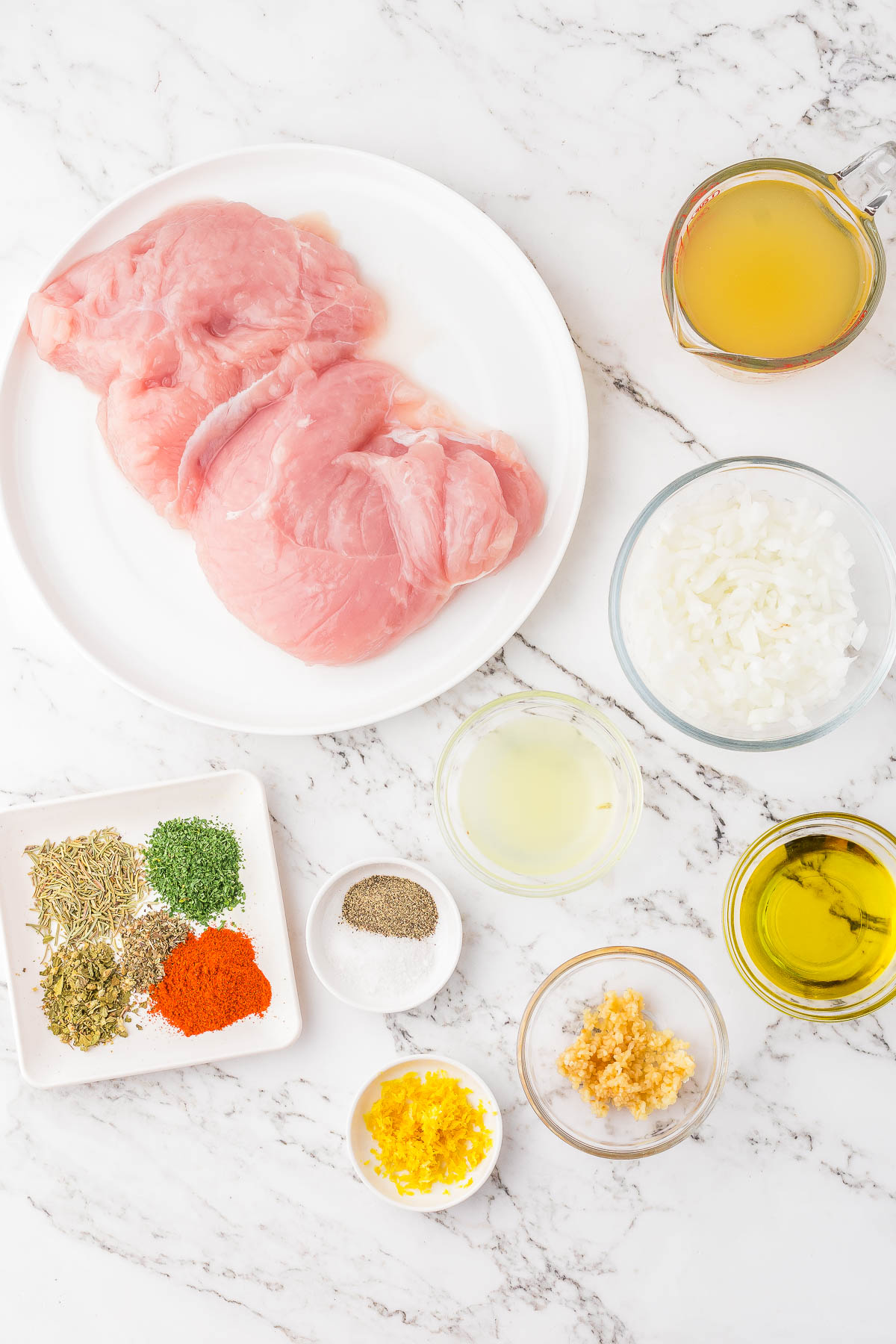 Raw chicken breasts on a plate, surrounded by bowls of chopped onions, spices, lemon zest, olive oil, chicken broth, minced garlic, and lemon juice on a marble surface.