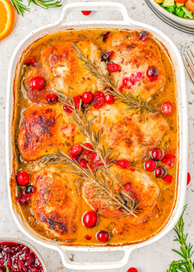 Baked chicken thighs with rosemary, cranberries, and orange slices in a white baking dish.